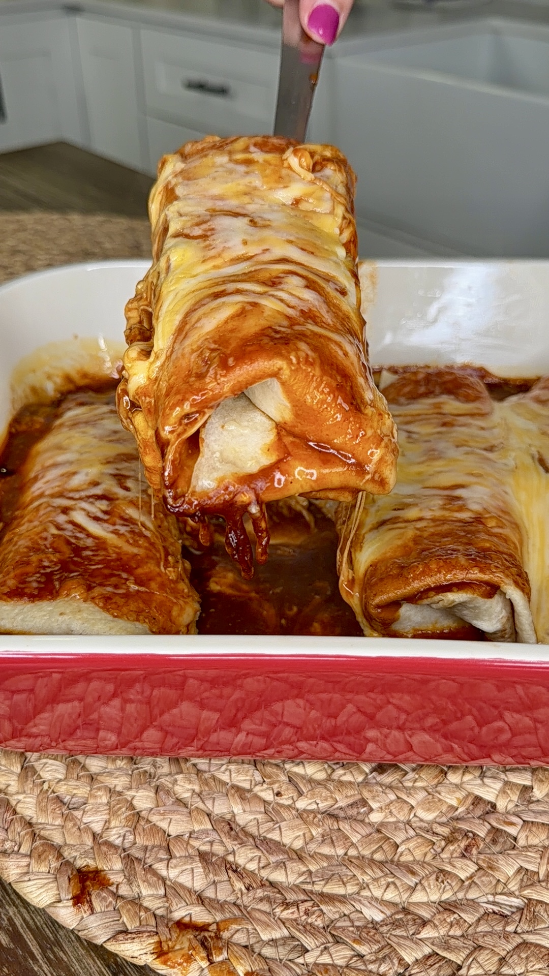 A hand lifts a cheesy, sauce-covered enchilada from a red baking dish filled with more enchiladas, sitting on a woven placemat in a kitchen setting.