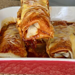A hand lifts a cheesy, sauce-covered enchilada from a red baking dish filled with more enchiladas. The dish sits on a woven mat in a kitchen setting.