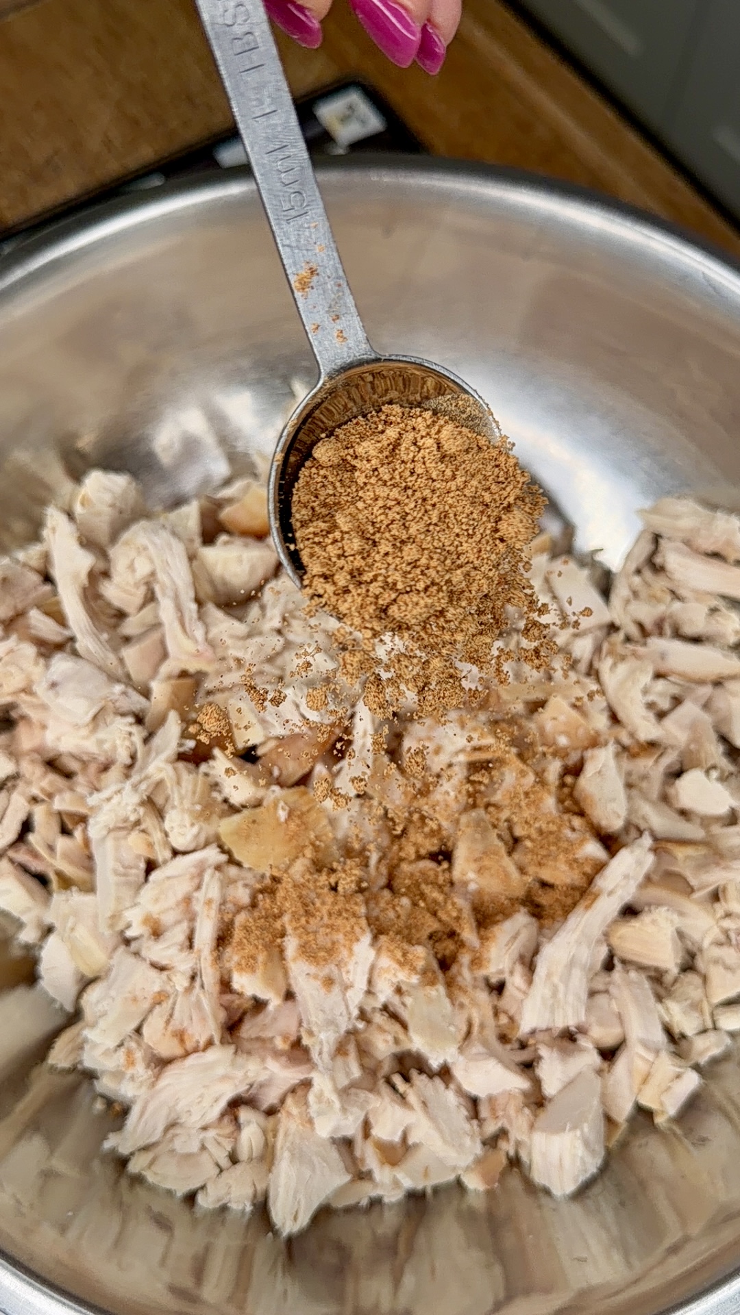 A metal tablespoon pours brown seasoning onto shredded chicken in a stainless steel mixing bowl. A hand with pink nails holds the spoon above the bowl.