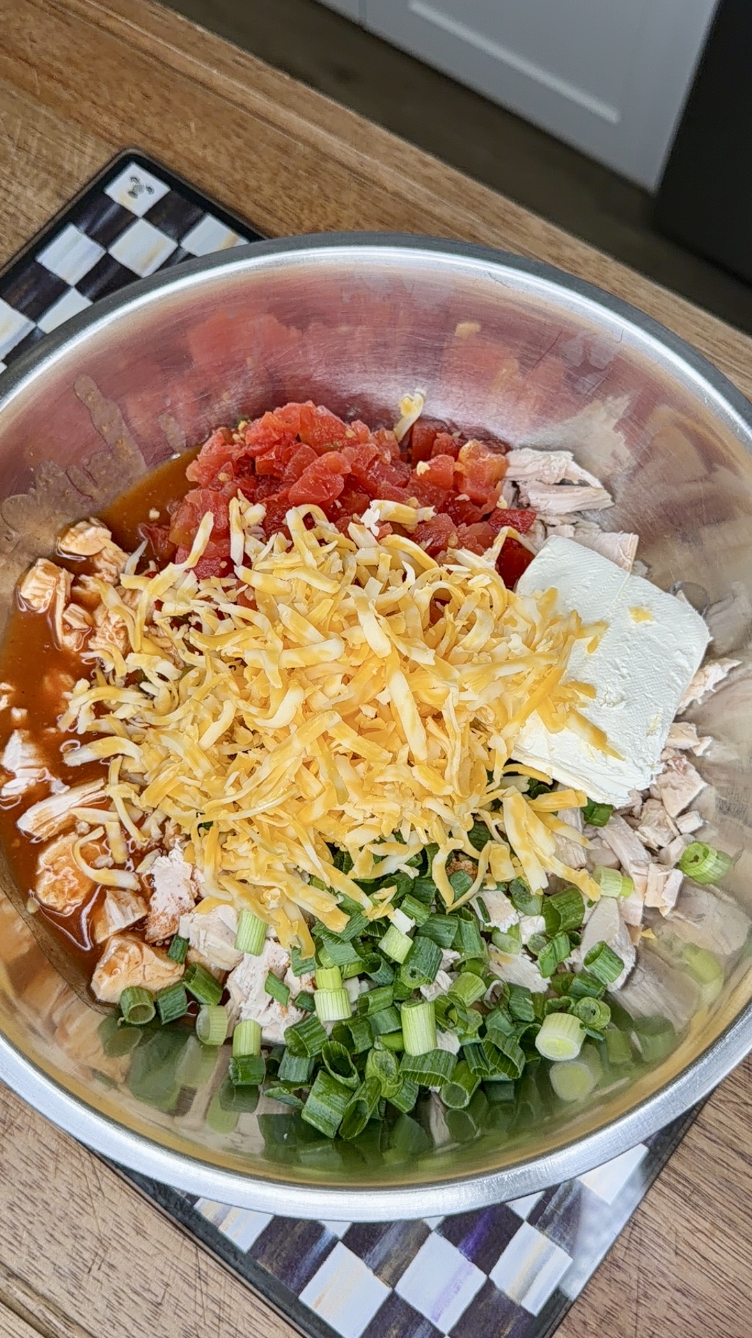 A metal mixing bowl on a wooden counter holds chopped green onions, shredded cheese, diced tomatoes, cubed cooked chicken, cream cheese, and a reddish sauce, all unmixed. A checkered trivet is partially visible beneath the bowl.