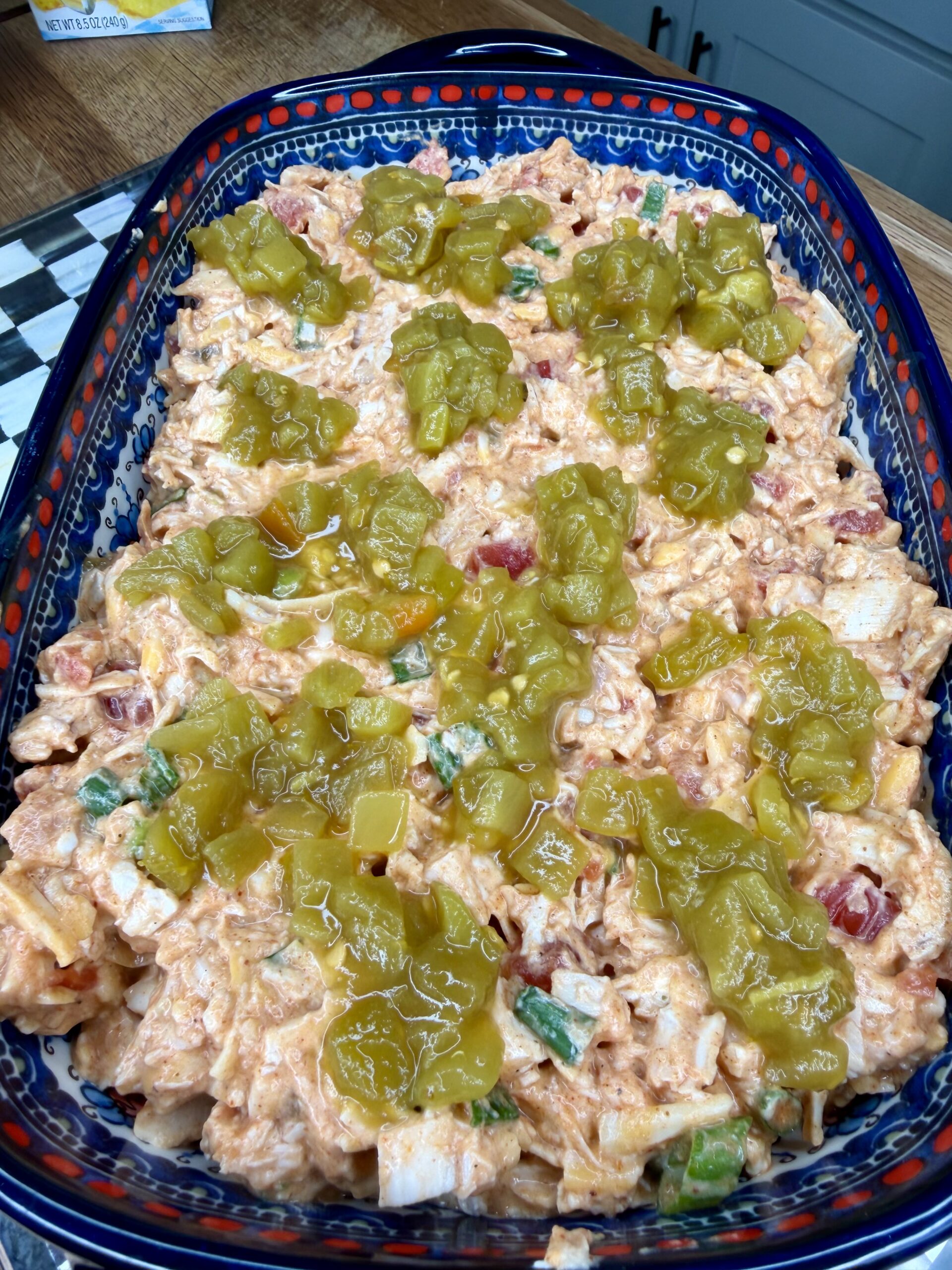 A casserole dish filled with a creamy mixture of shredded chicken, diced vegetables, and sauce, topped with dollops of chopped green chiles. The dish is ready for baking, sitting on a kitchen countertop.