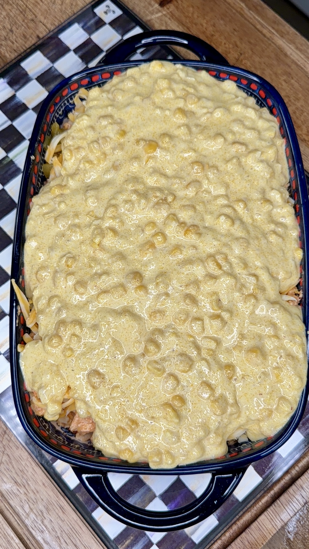 A rectangular baking dish filled with uncooked macaroni and cheese, topped with a thick, bubbly cheese sauce, sits on a checkered placemat on a wooden surface.