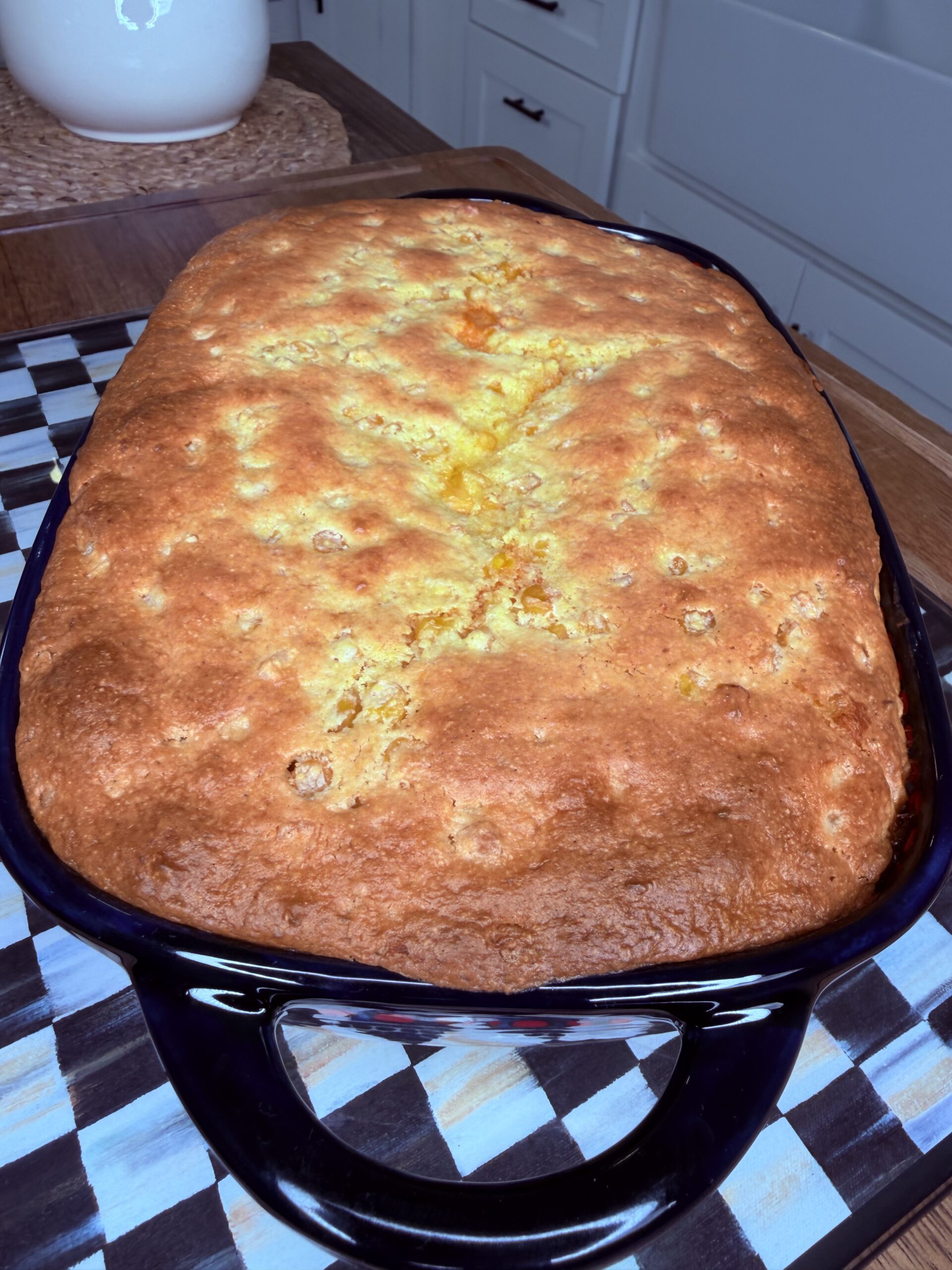 A golden-brown baked casserole sits in a dark baking dish on a checkered trivet, on a wooden kitchen counter with white cabinets in the background.