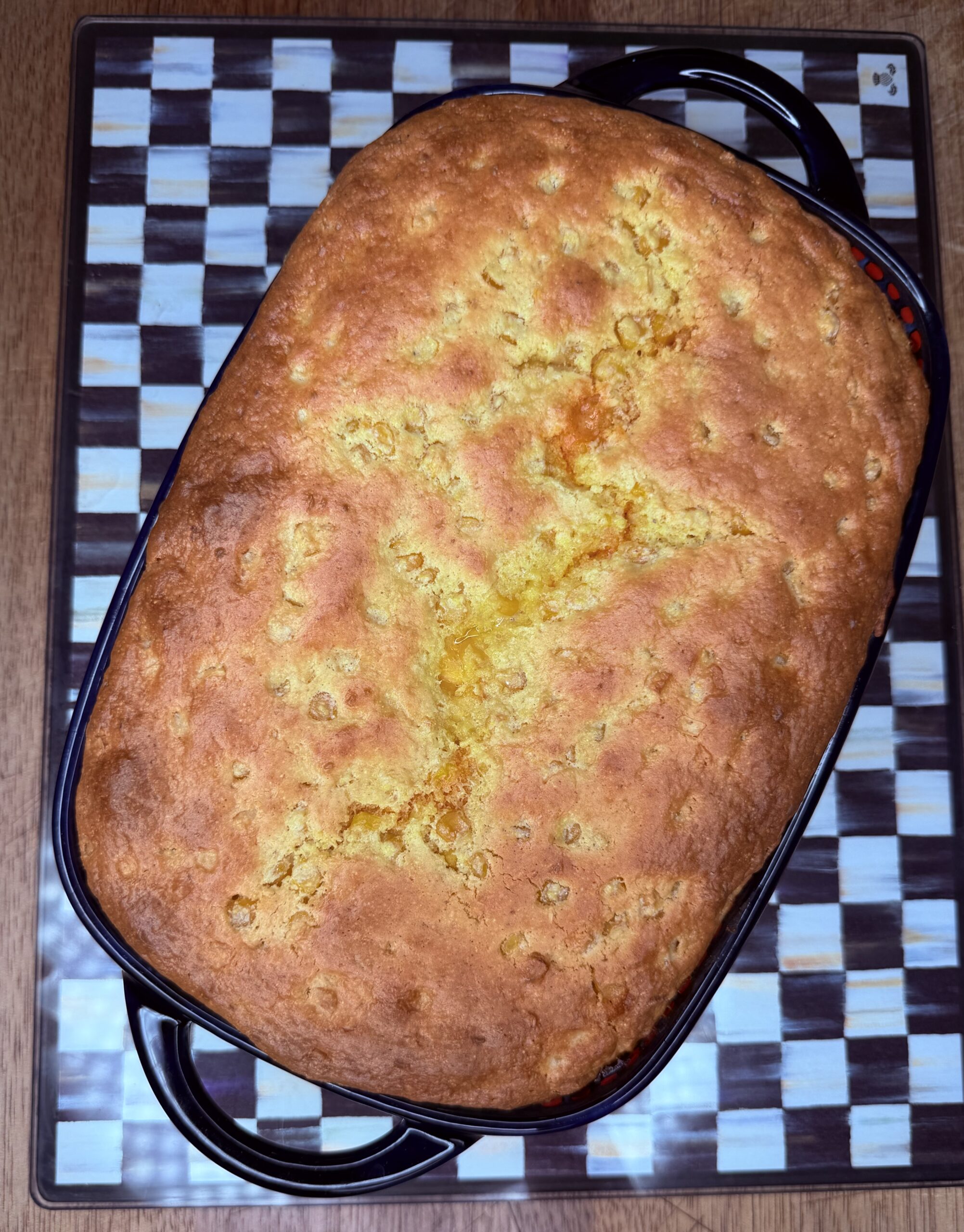 A golden-brown baked cornbread sits in a rectangular black baking dish on a checkered-pattern tray atop a wooden table.