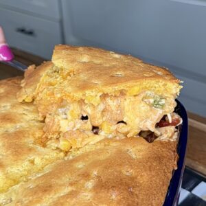 A hand with pink nail polish lifts a square serving of golden-brown cornbread casserole from a baking dish, revealing a cheesy filling with corn, beans, and vegetables.