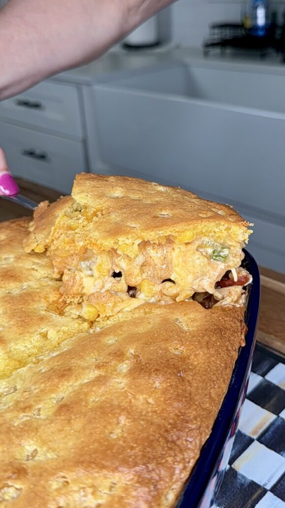 A hand with pink nail polish lifts a square serving of golden-brown cornbread casserole from a baking dish, revealing a cheesy filling with corn, beans, and vegetables.