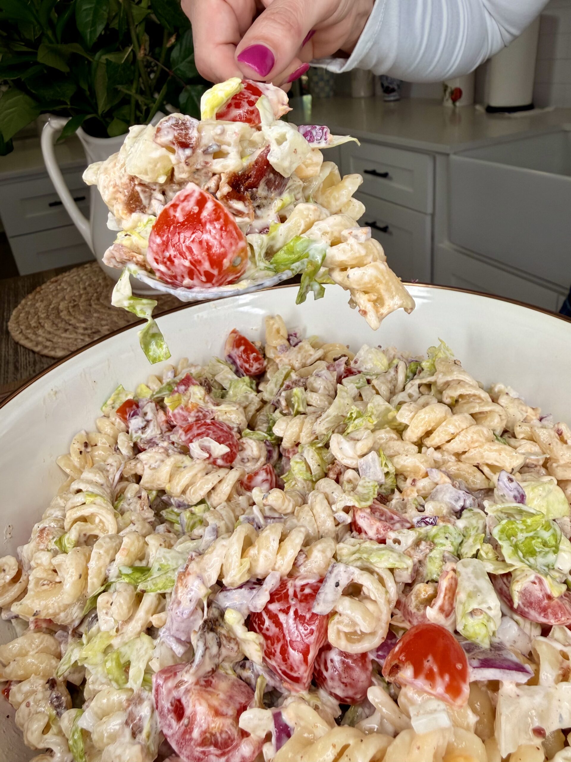 A hand holds a serving spoonful of creamy pasta salad with rotini, cherry tomatoes, lettuce, and bacon over a large bowl of the same salad in a kitchen setting.