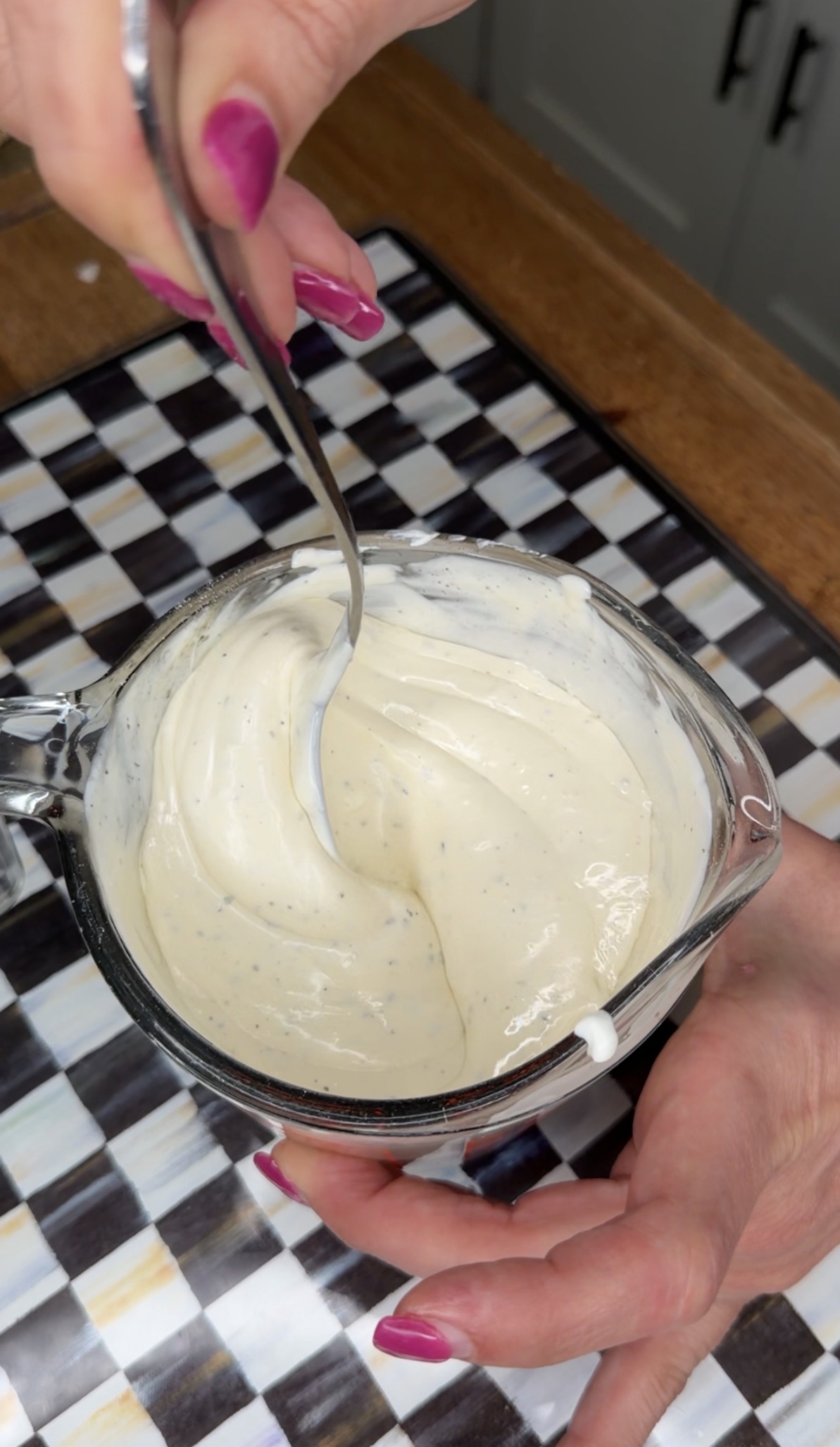A hand holds a glass measuring cup filled with creamy, speckled sauce while another hand stirs it with a spoon over a black-and-white checkered surface.