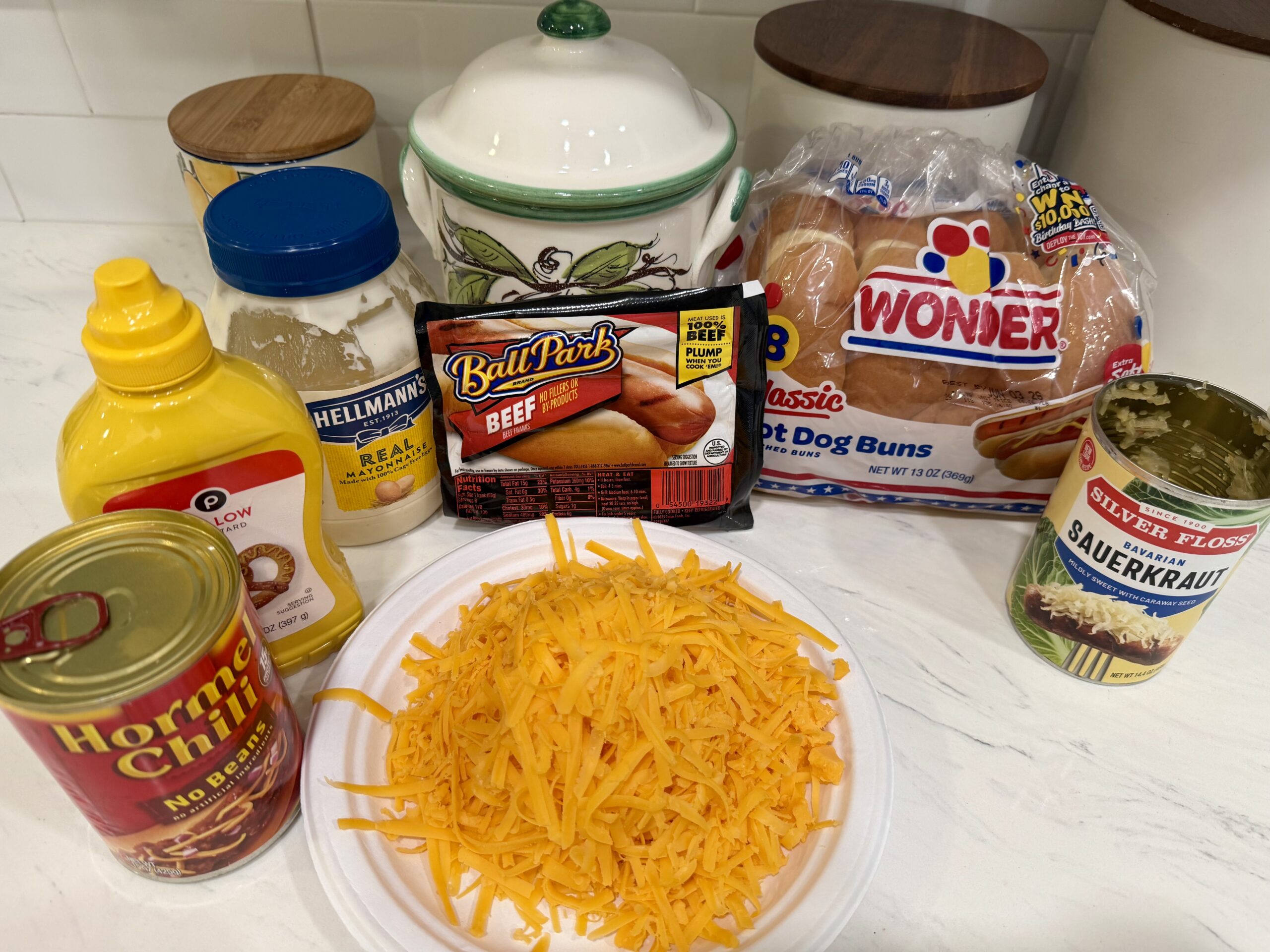 A kitchen counter displays ingredients for The Ultimate Hotdog Bake: mustard, mayonnaise, canned chili, shredded cheddar cheese, beef hot dogs, hot dog buns, and sauerkraut, with a ceramic jar in the background.