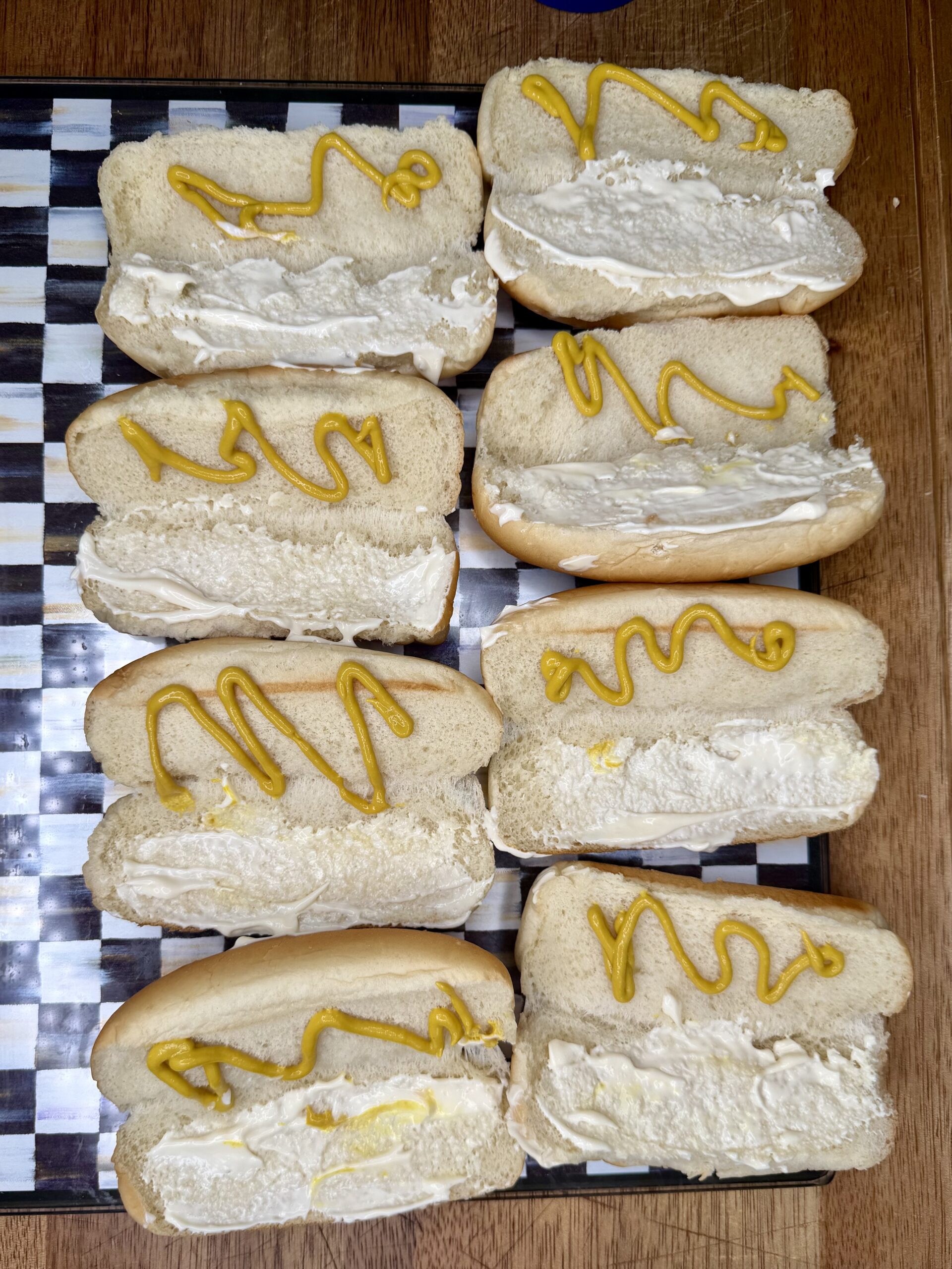 Eight hot dog buns arranged on a checkered tray, each spread with mayonnaise and topped with a squiggle of yellow mustard; this is The Ultimate Hotdog Bake—though there are no sausages present in the buns.