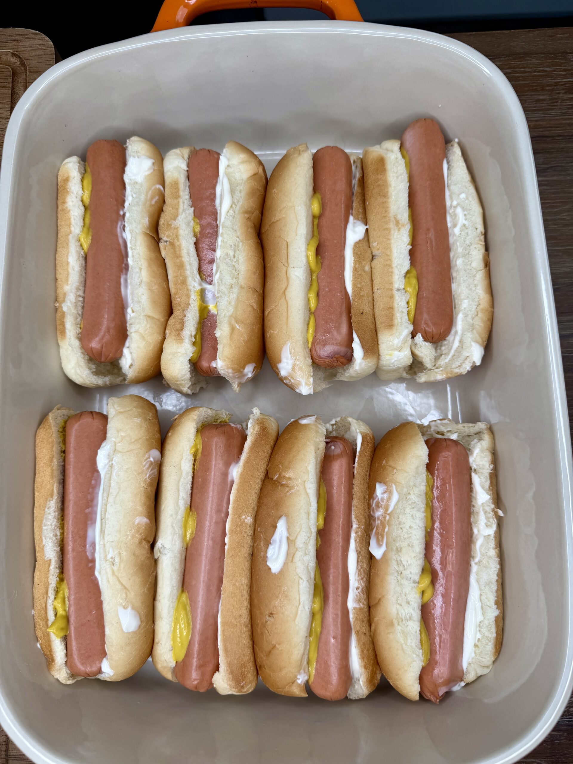Eight hot dogs in buns are arranged in a rectangular baking dish for The Ultimate Hotdog Bake. Some buns have mayonnaise, some mustard, and others both condiments. The hot dogs are ready to be served or cooked further.