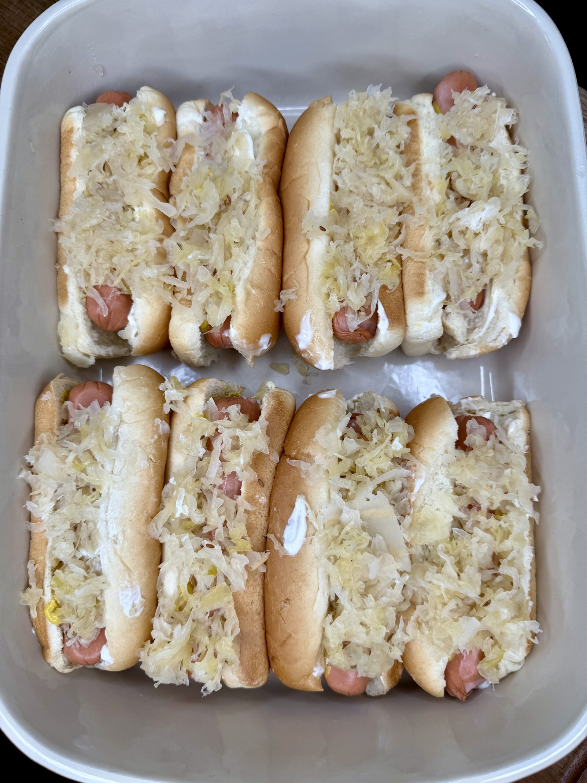 A container holds eight hot dogs in buns, each topped with sauerkraut and a visible layer of mayonnaise or a similar spread. The hot dogs, arranged neatly in two rows, make for The Ultimate Hotdog Bake experience.