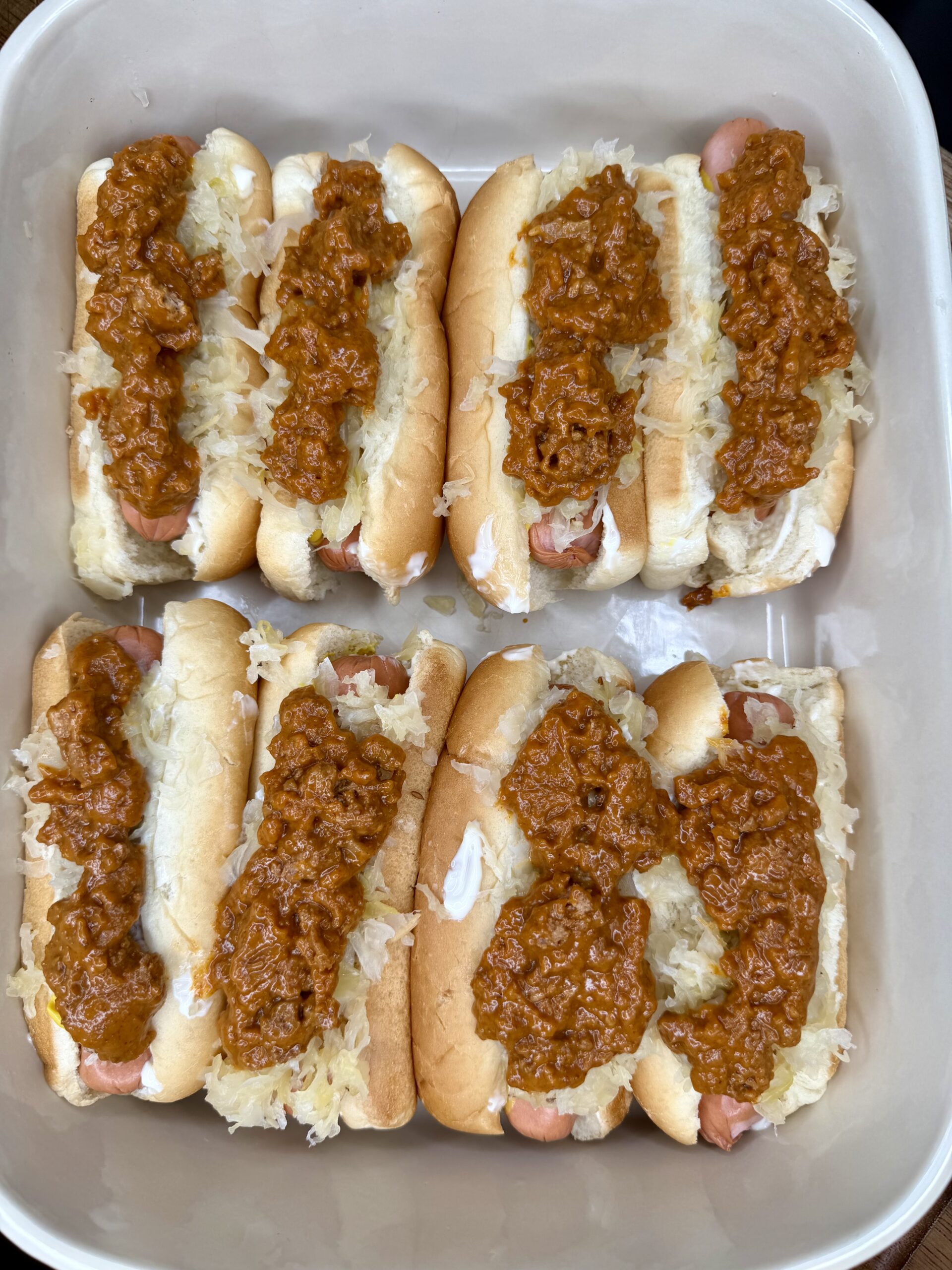 Six hot dogs in buns topped with sauerkraut and chili are arranged in a baking dish. The Ultimate Hotdog Bake features each hot dog neatly lined up, generously covered with sauerkraut and a layer of hearty chili.