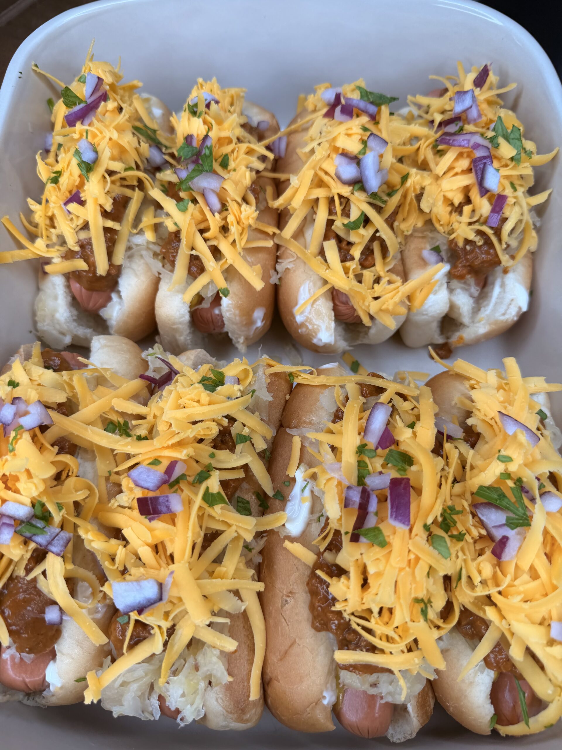 The Ultimate Hotdog Bake features six hot dogs in buns, topped with shredded cheddar cheese, chopped red onions, and fresh herbs in a white dish. Chili and other condiments are layered beneath the cheese for extra flavor.