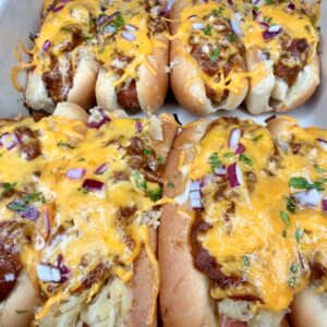 The Ultimate Hotdog Bake: Six hot dogs in buns topped with melted cheddar cheese, chili, chopped red onions, fresh herbs, and a touch of sauerkraut, all arranged closely together in a white baking dish.