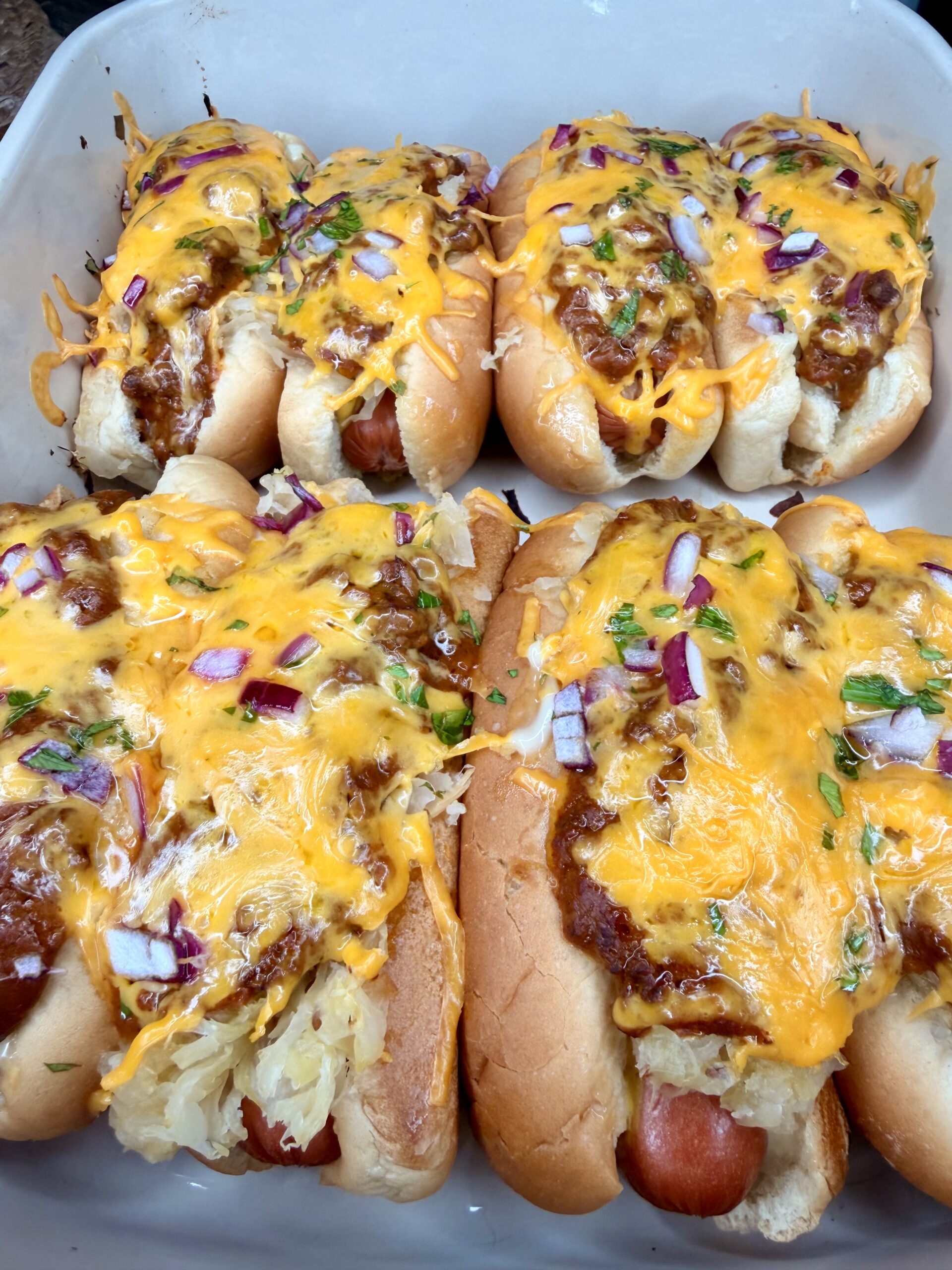 The Ultimate Hotdog Bake: Six hot dogs in buns topped with melted cheddar cheese, chili, chopped red onions, fresh herbs, and a touch of sauerkraut, all arranged closely together in a white baking dish.