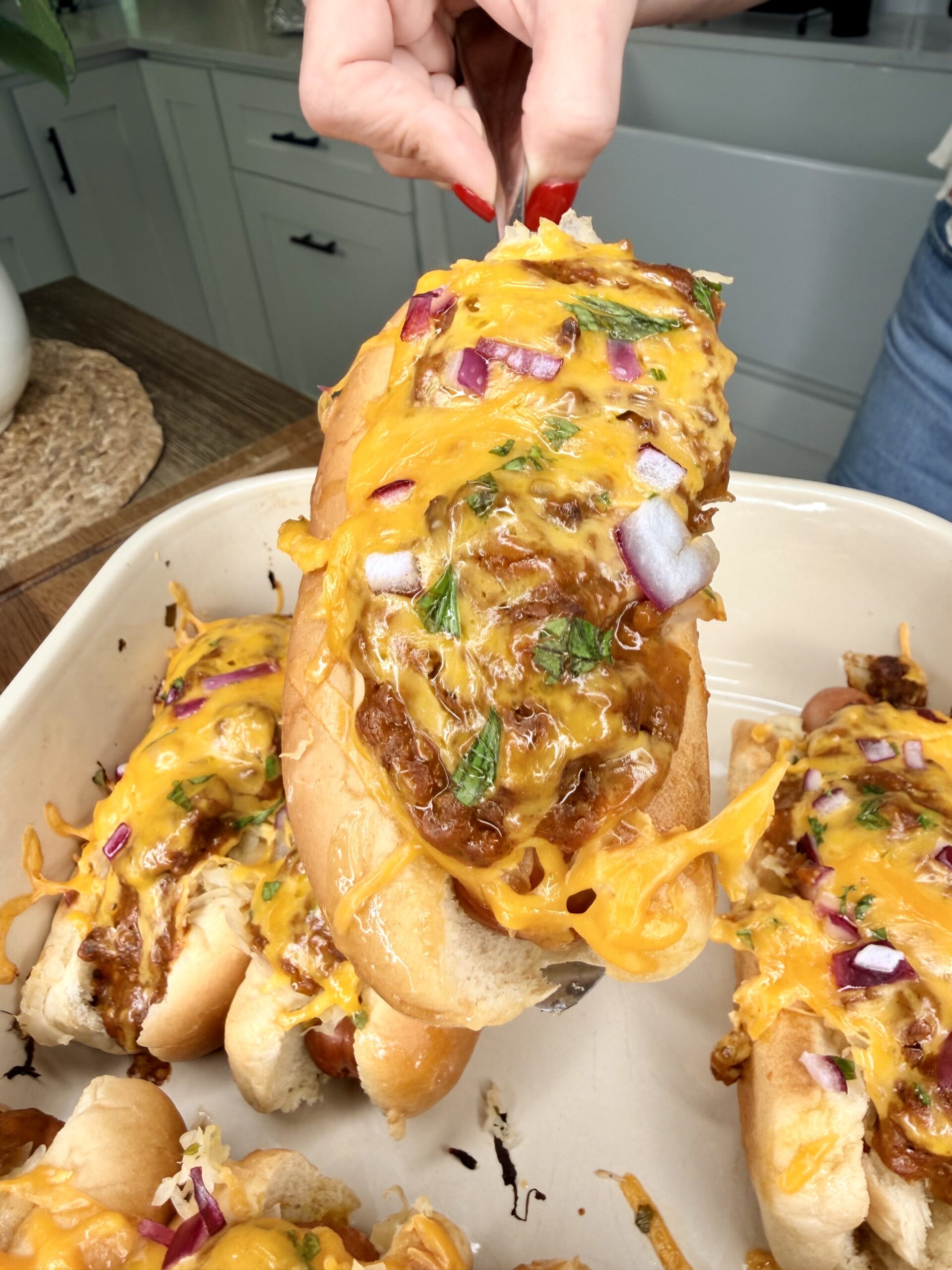 A hand lifts a chili cheese hot dog from a baking dish. The Ultimate Hotdog Bake features hot dogs topped with melted cheddar, chopped red onions, and fresh herbs; other deliciously loaded hot dogs are visible below.