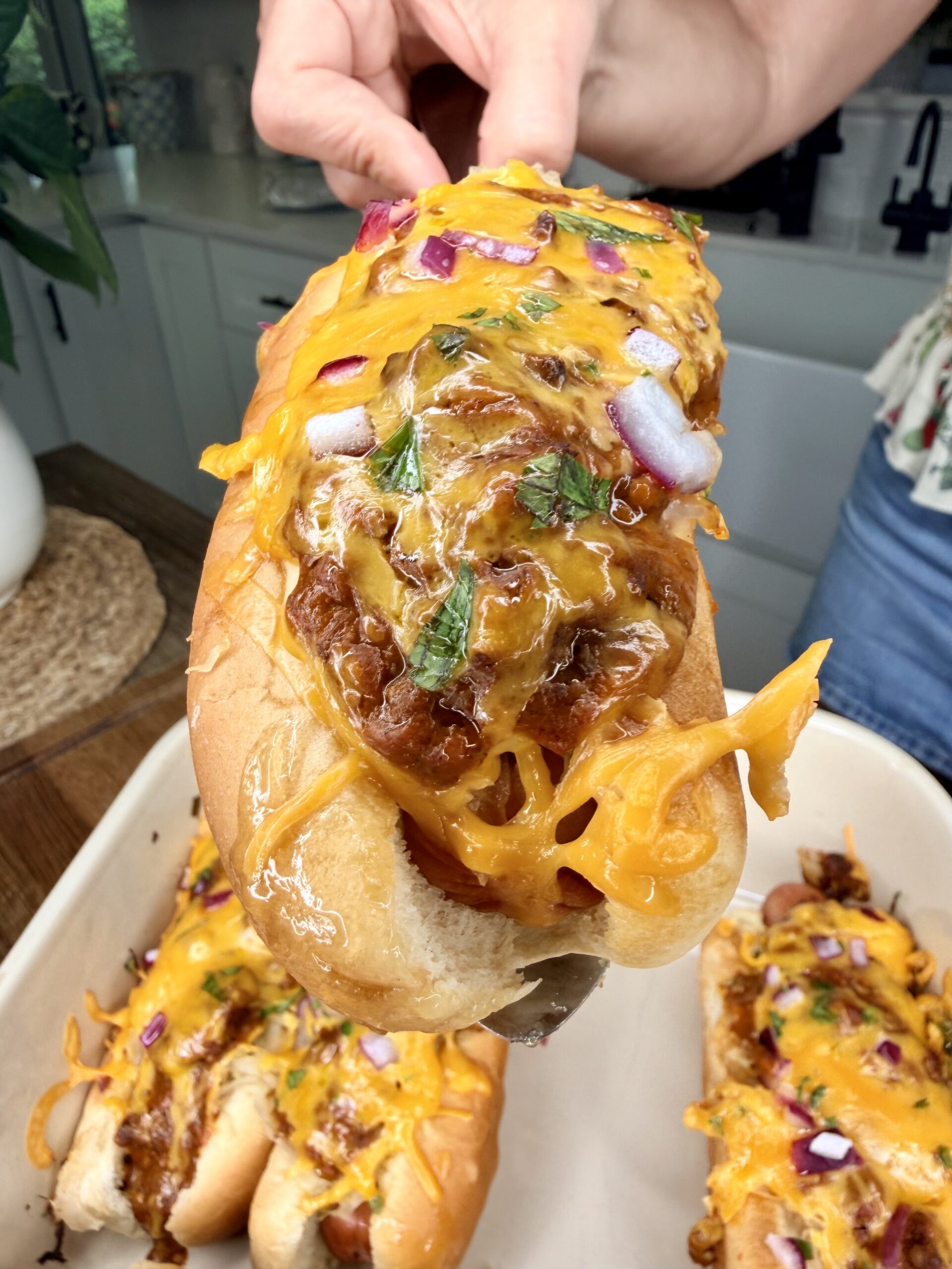A close-up of a hand holding The Ultimate Hotdog Bake—a chili cheese hot dog topped with melted cheddar, chopped red onions, and herbs, with more hot dogs in a dish below in a cozy kitchen setting.