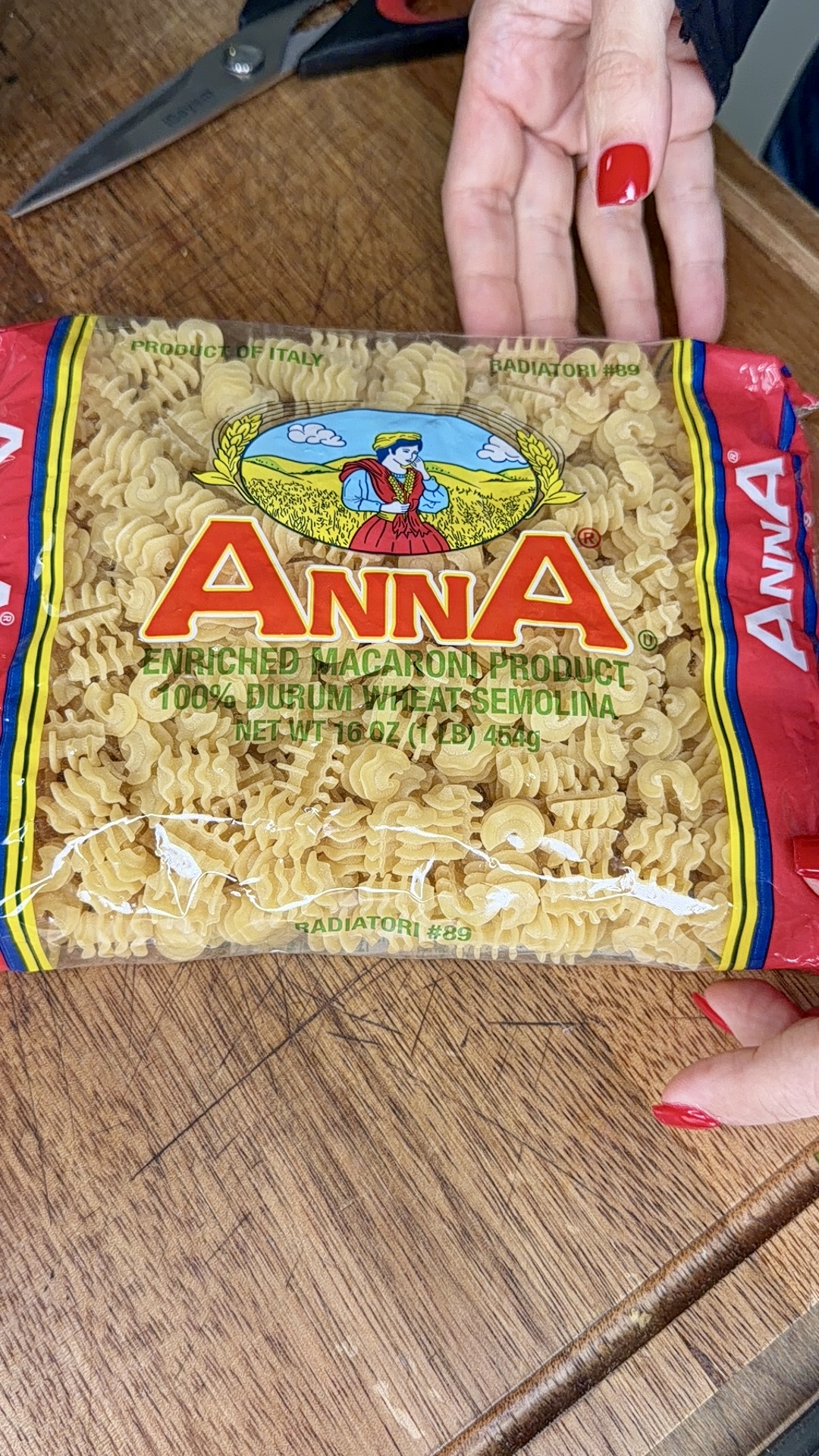 A person with red nail polish holds a package of Anna brand rotini pasta labeled Enriched Macaroni Product, 100% Durum Wheat Semolina, Net Wt. 16 oz (1 lb) 454 g on a wooden surface with scissors nearby.