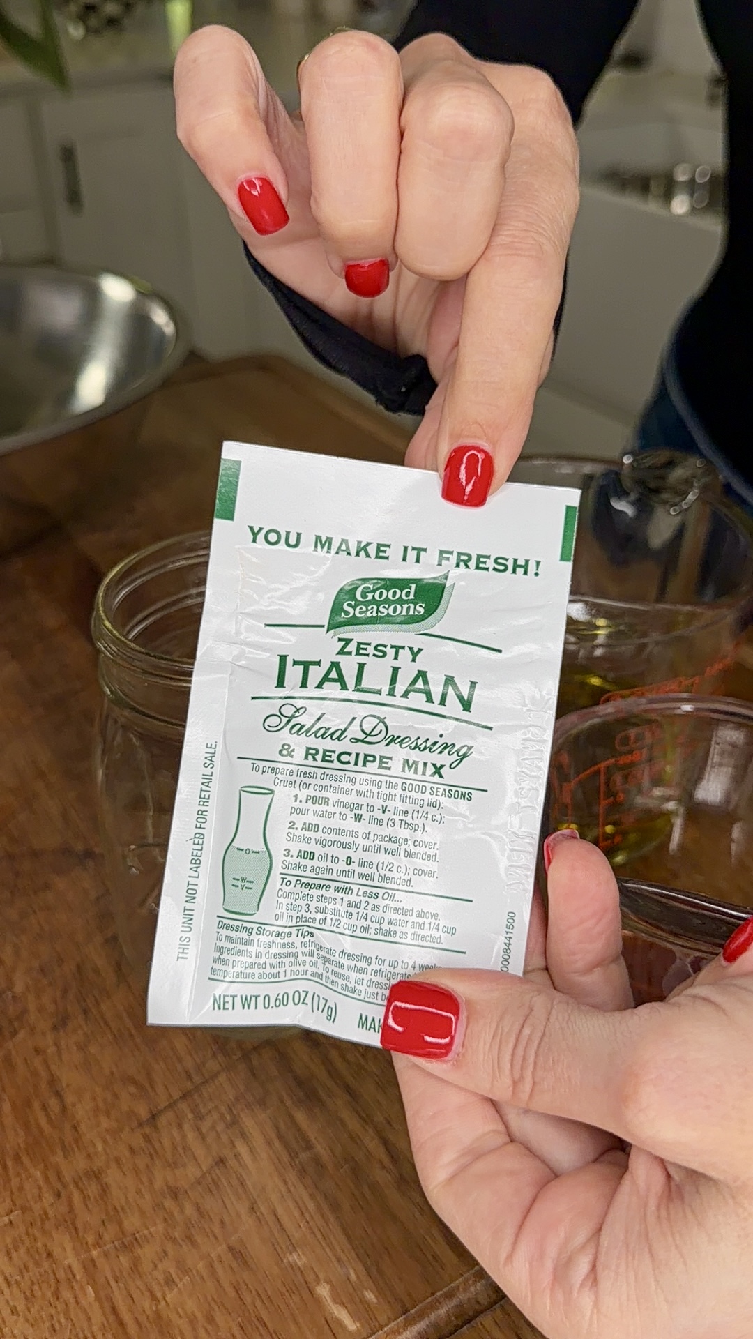 A person with red nail polish holds a packet of Good Seasons Zesty Italian Salad Dressing & Recipe Mix above a glass container on a wooden countertop.