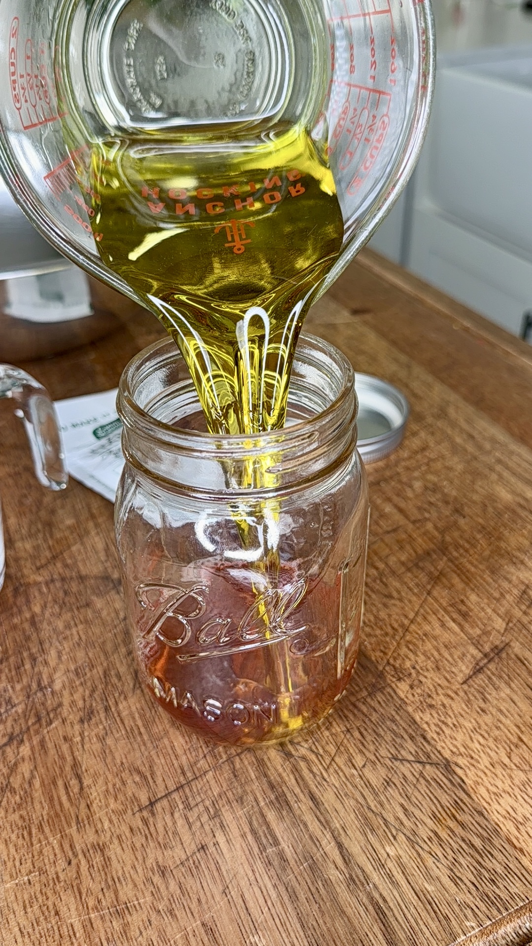A glass measuring cup pours golden oil into a clear Ball mason jar that already contains a small amount of oil, all set on a wooden countertop with kitchen items in the background.