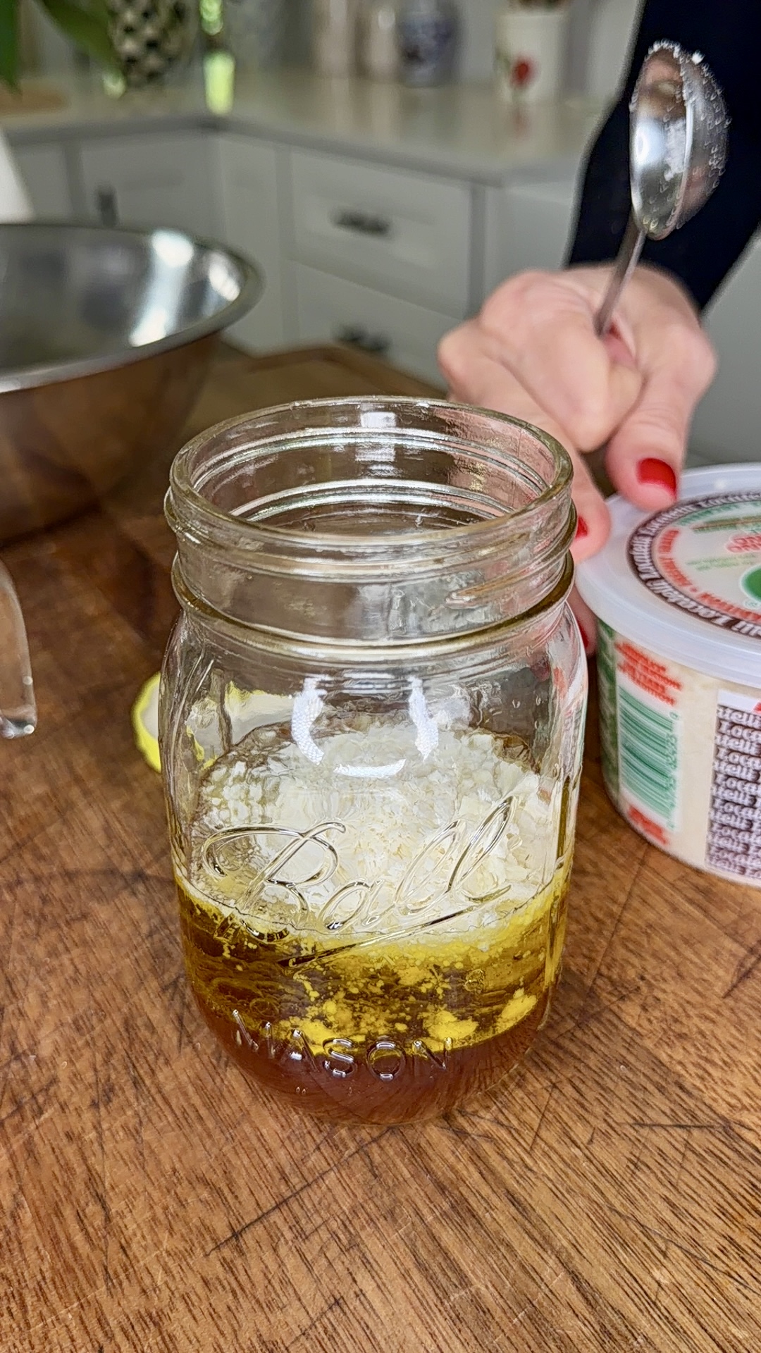 A glass jar on a wooden countertop contains oil and seasonings. Behind it, a hand with red nails holds a spoon above a tub of grated cheese, with a metal mixing bowl nearby. Kitchen items are visible in the background.