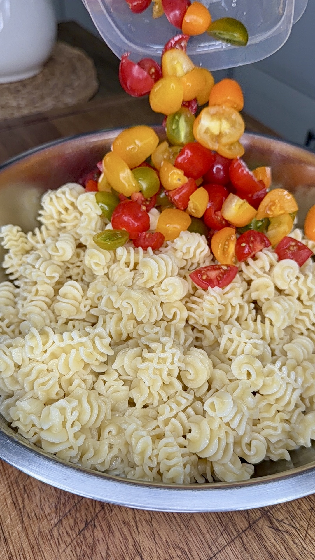 A metal bowl filled with cooked radiatori pasta, with a hand pouring colorful chopped cherry tomatoes from a container on top, set on a wooden surface.
