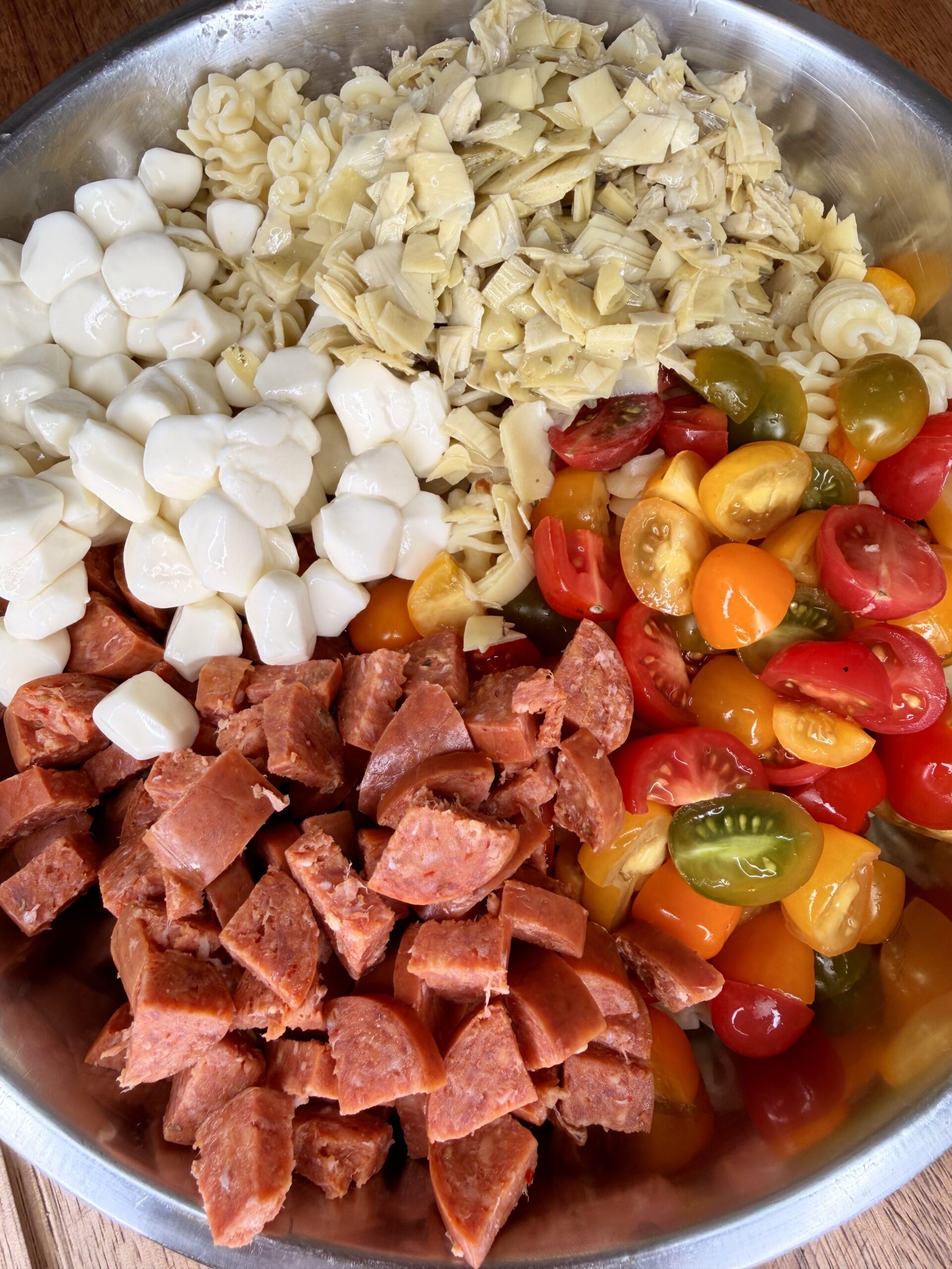 A bowl filled with separated ingredients: mozzarella balls, chopped artichoke hearts, halved cherry tomatoes in various colors, sliced pepperoni, and cooked pasta.