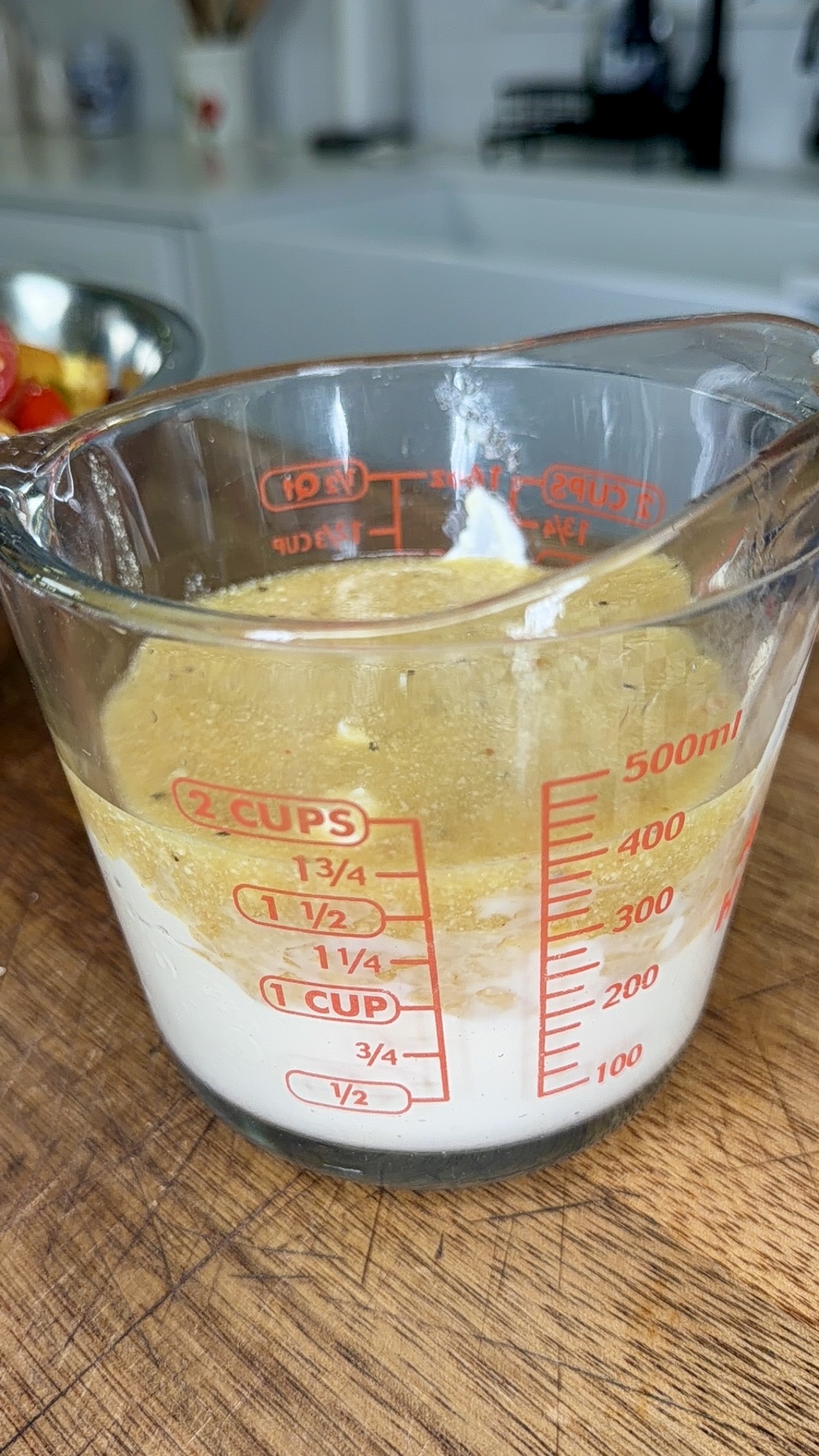 A glass measuring cup containing flour, a thick tan liquid, and a dollop of white substance sits on a wooden counter in a kitchen. The cup displays red measurement markings up to 2 cups or 500 milliliters.