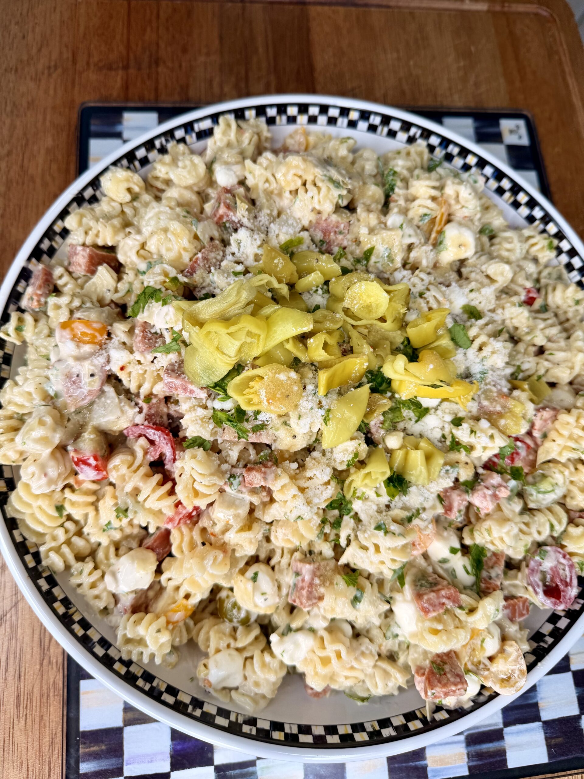 A large bowl of pasta salad with rotini, chopped vegetables, herbs, grated cheese, and sliced yellow pepperoncini, served on a patterned plate atop a wooden table.