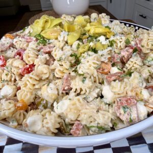 A large white bowl filled with creamy rotini pasta salad mixed with mozzarella balls, tomatoes, herbs, salami, and sliced pepperoncini, sitting on a checkered cloth in a kitchen setting.