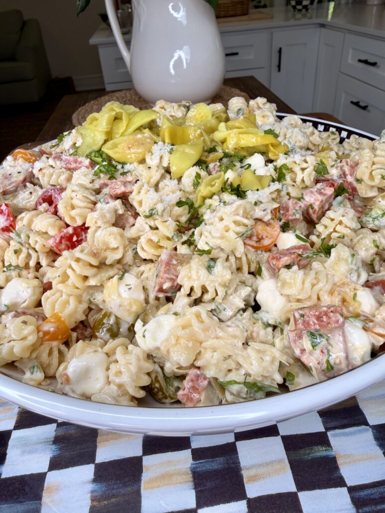 A large white bowl filled with creamy rotini pasta salad mixed with mozzarella balls, tomatoes, herbs, salami, and sliced pepperoncini, sitting on a checkered cloth in a kitchen setting.