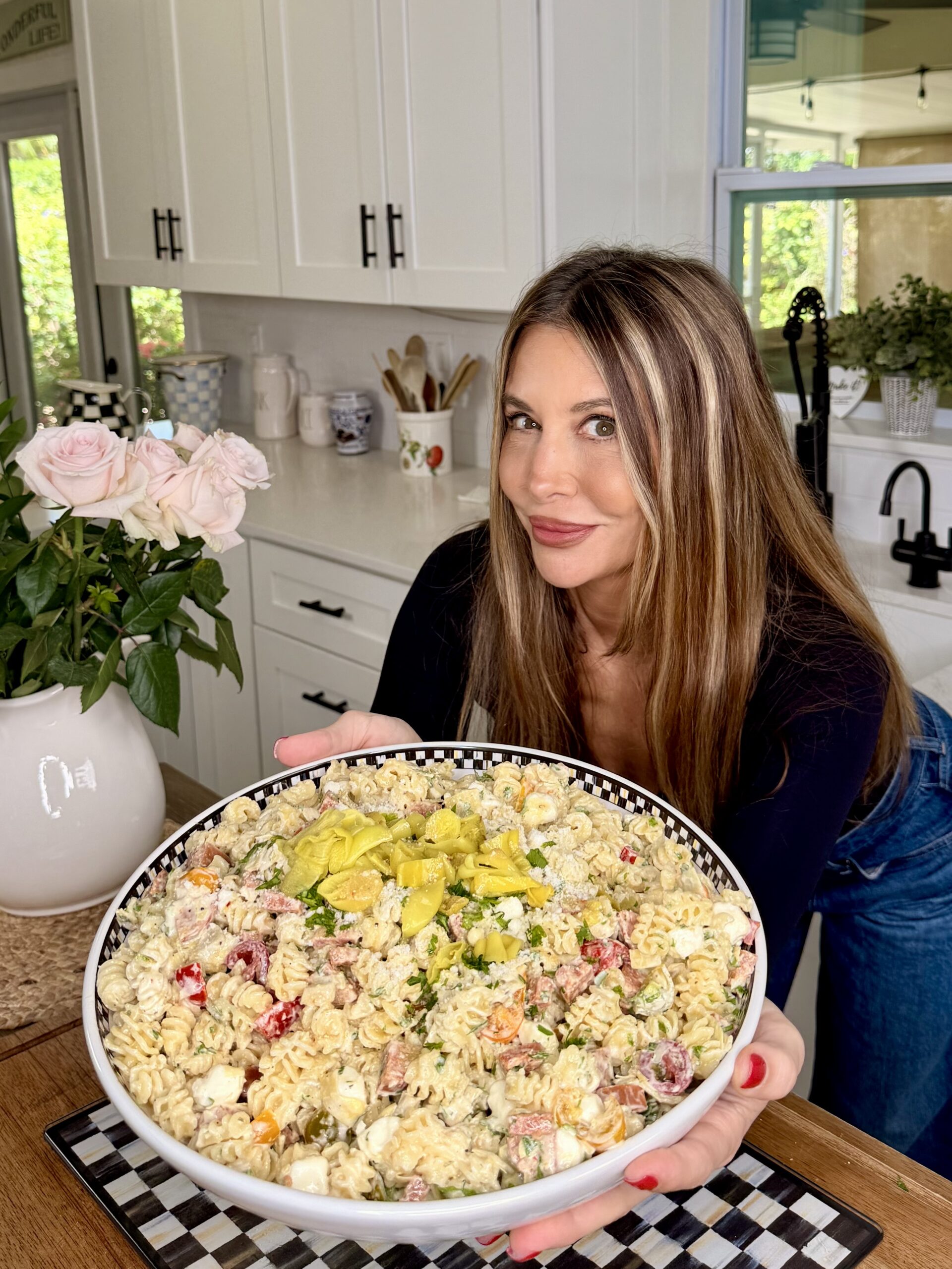 A woman with long brown hair, wearing a black top, smiles and holds a large bowl of pasta salad in a bright kitchen with white cabinets and a vase of pink roses on the counter.