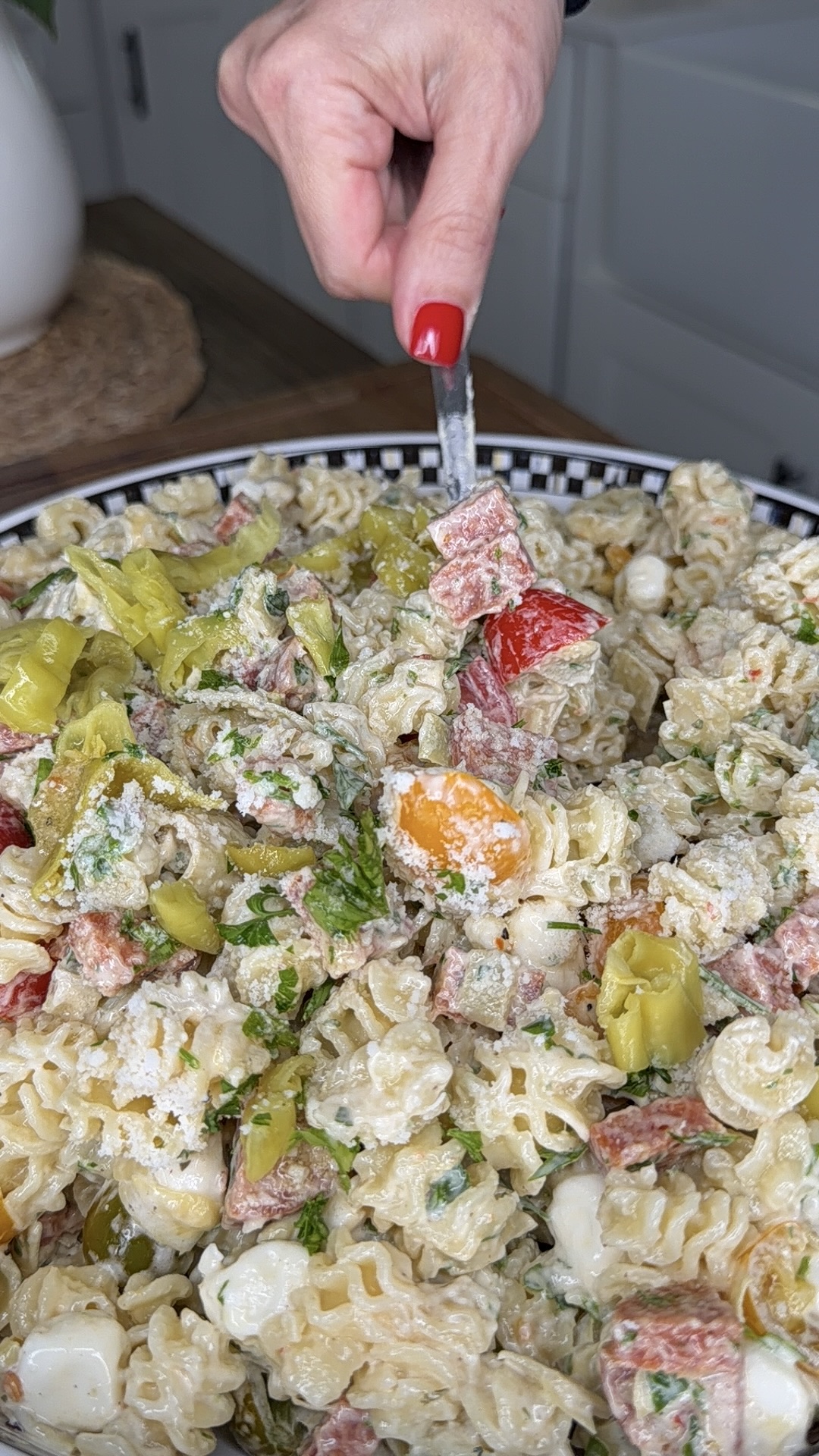 A hand with red nail polish holds a fork, mixing a creamy pasta salad with curly noodles, sliced peppers, mozzarella balls, herbs, and salami pieces in a large bowl.