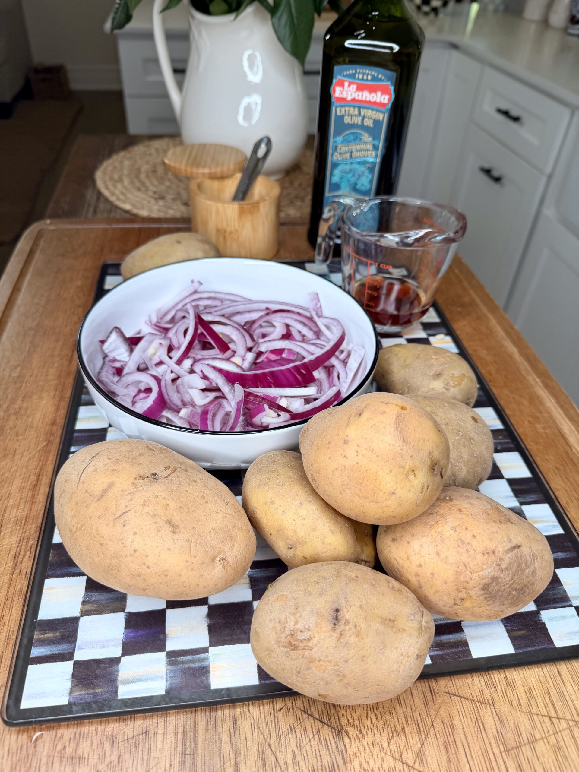 A bowl of sliced red onions sits on a checkered cutting board next to several whole potatoes, a measuring cup with liquid, a bottle of olive oil, and a wooden container with a spoon on a kitchen counter.