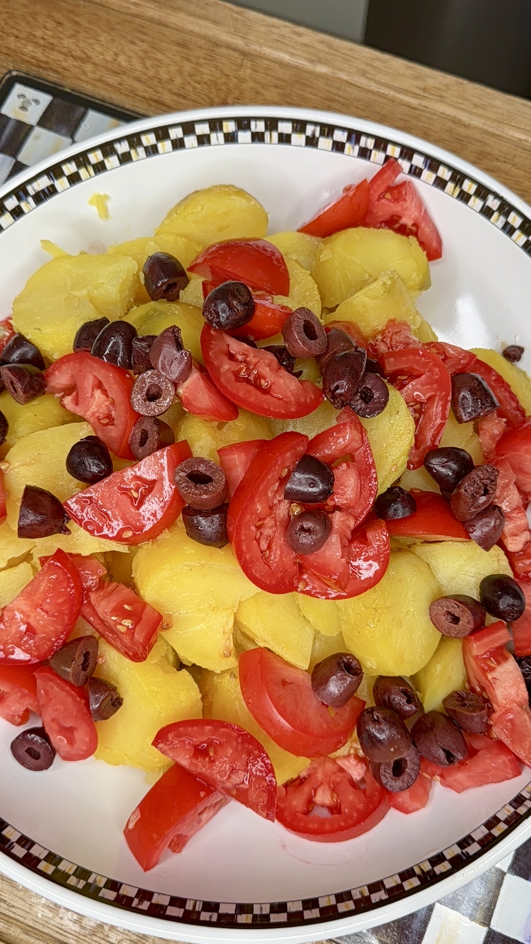 Sliced boiled potatoes topped with chopped fresh tomatoes and sliced black olives, served on a white plate with a checkered rim. The colorful salad rests on a patterned placemat.