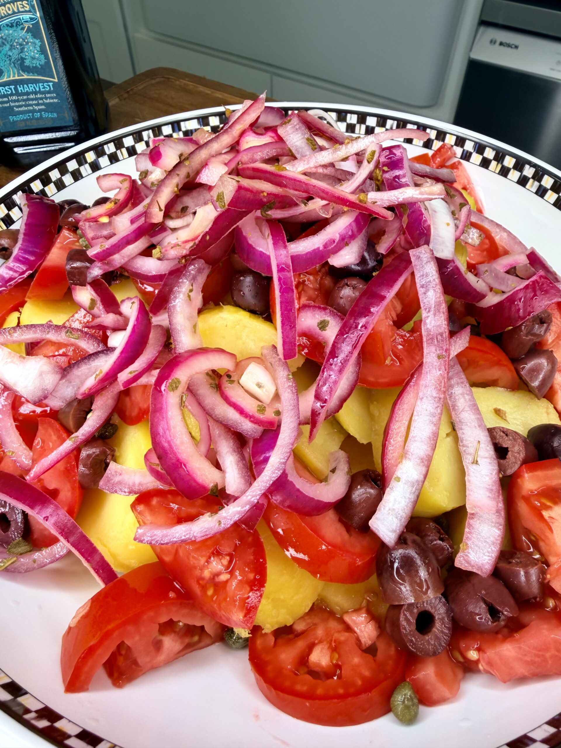 A close-up of a salad with sliced tomatoes, yellow potatoes, kalamata olives, capers, and thinly sliced red onions, served in a patterned bowl on a kitchen counter.