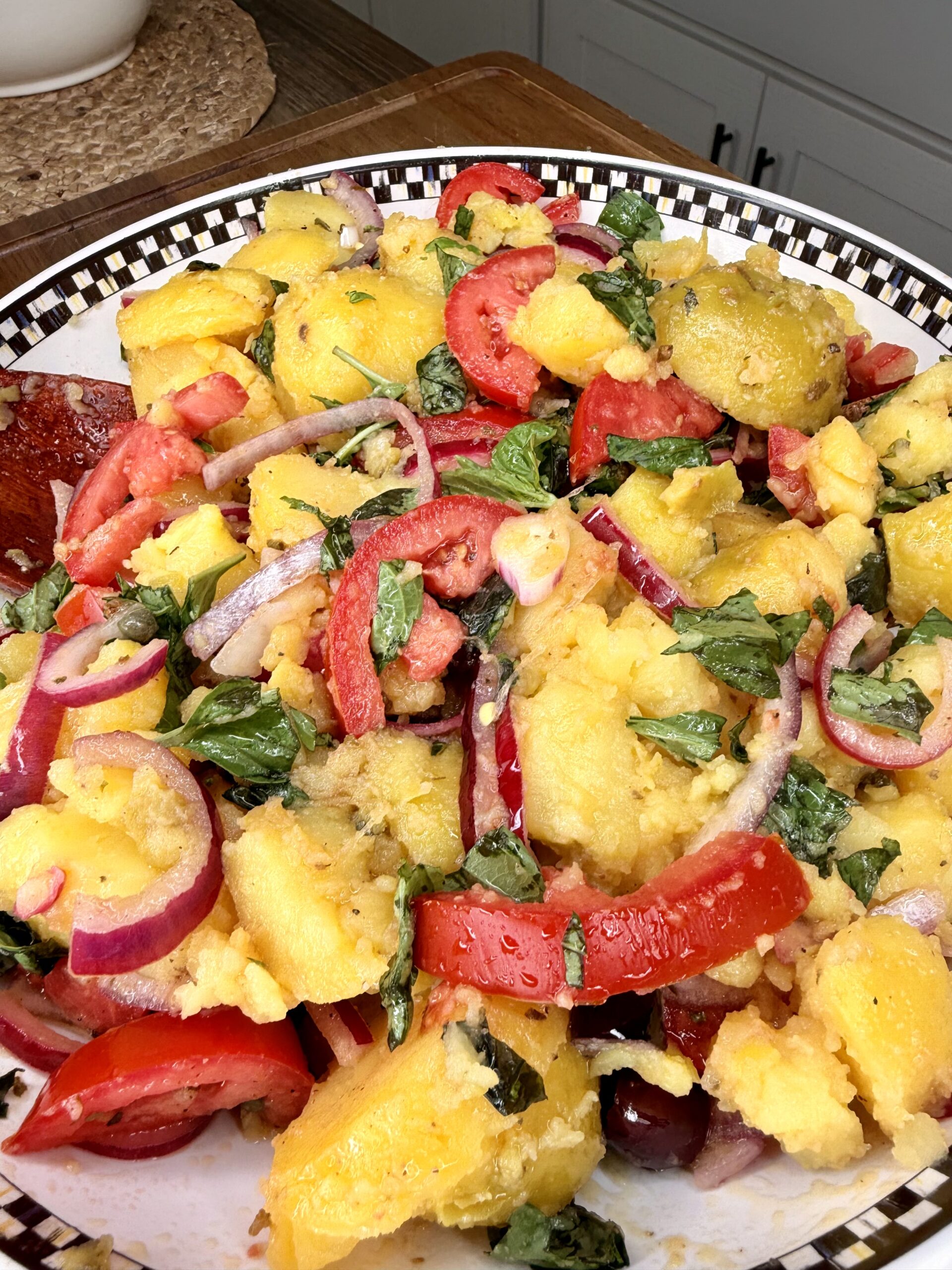 A close-up of a plate with a colorful salad made of yellow potatoes, tomato slices, red onion, chopped fresh basil, and a light vinaigrette, served on a checkered rim dish.