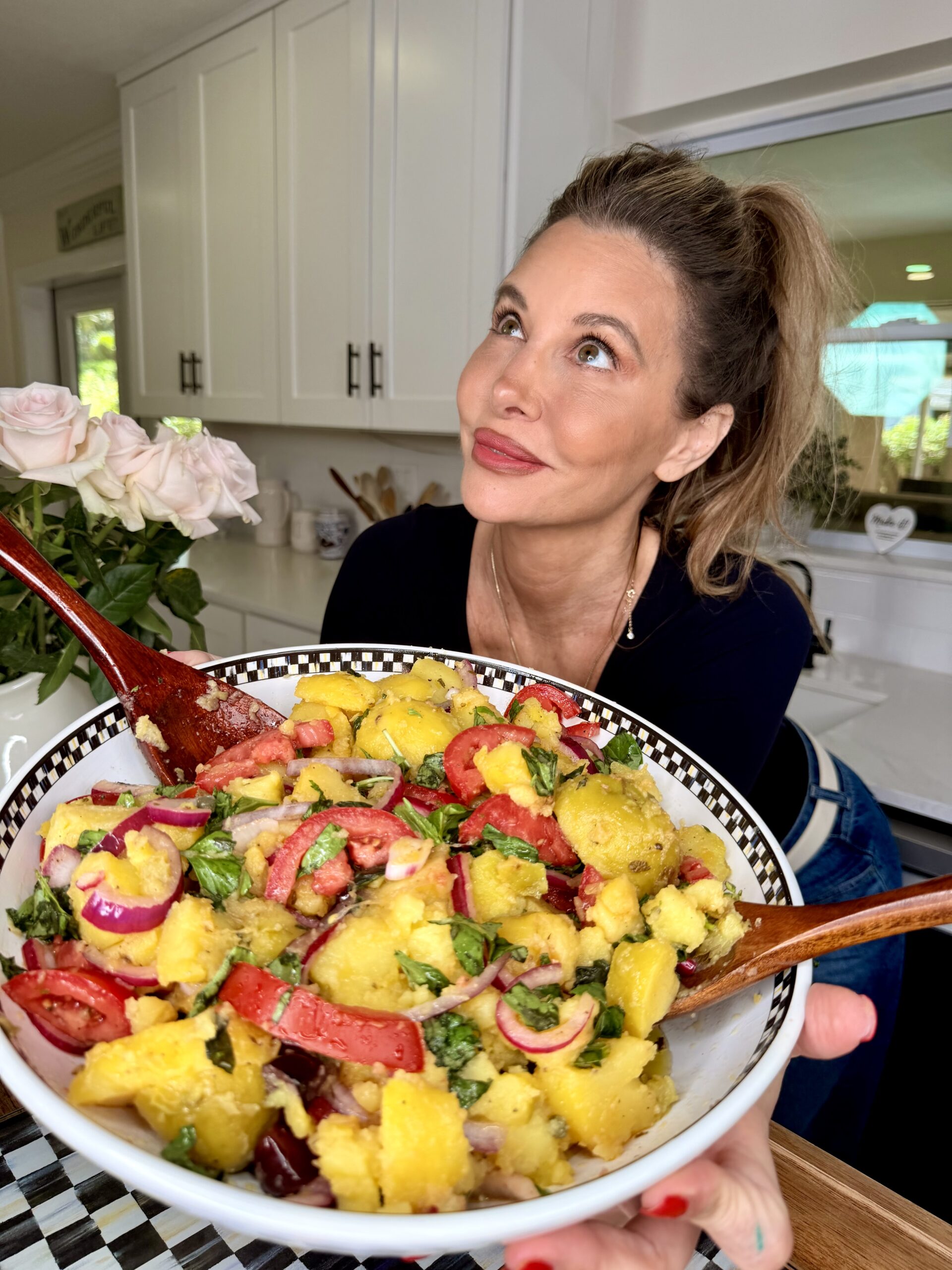 A woman in a kitchen holds a large bowl of colorful salad with tomatoes, onions, basil, and bread pieces, smiling and looking upward.