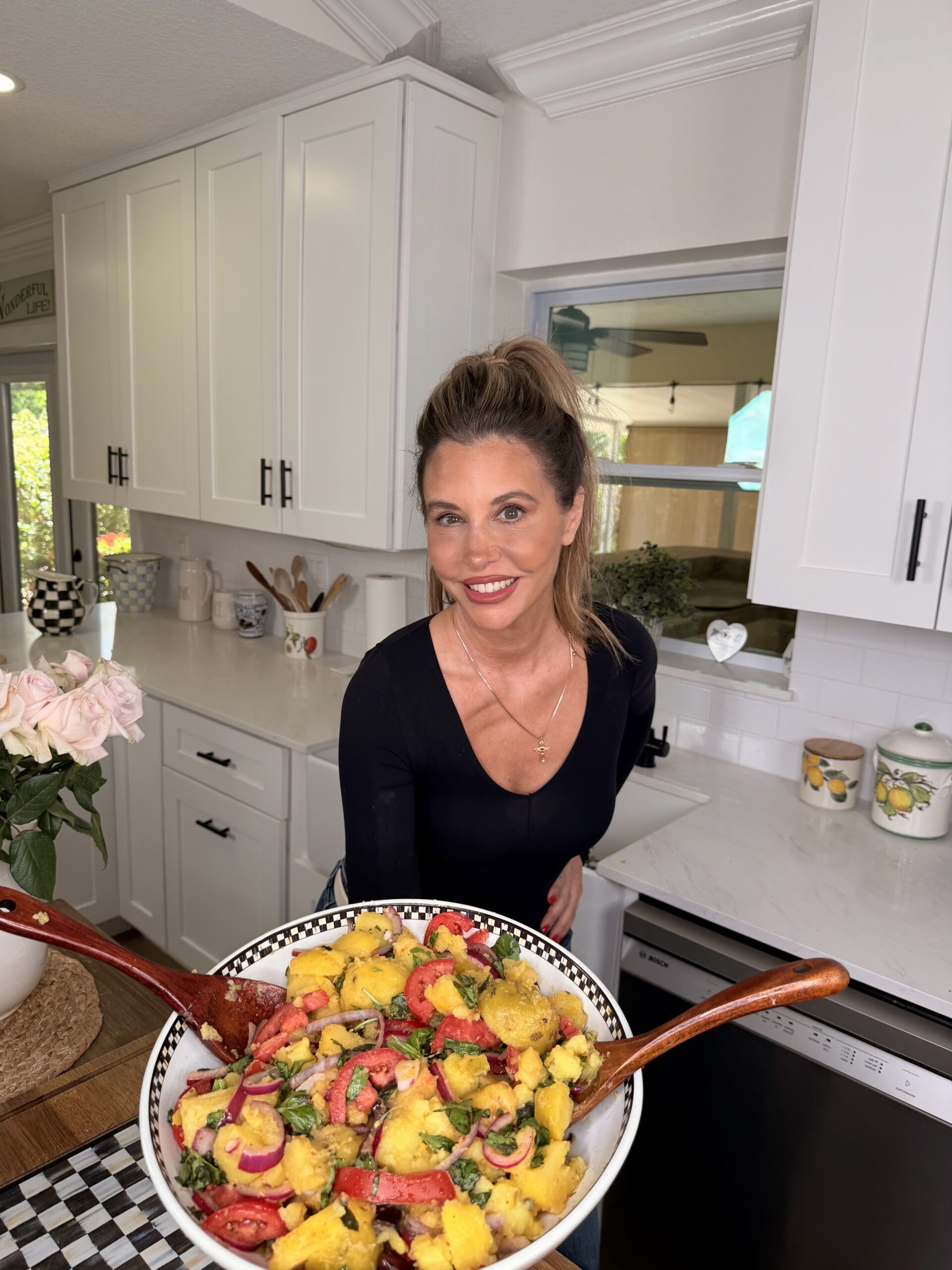 A woman with long hair in a black top stands in a bright kitchen, smiling at the camera, holding a large bowl of colorful salad with wooden utensils. White cabinets and flowers are visible in the background.