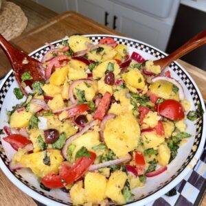 A bowl of vibrant potato salad with sliced red onions, tomatoes, olives, chopped herbs, and red peppers, mixed with a fork and spoon, set on a kitchen counter.