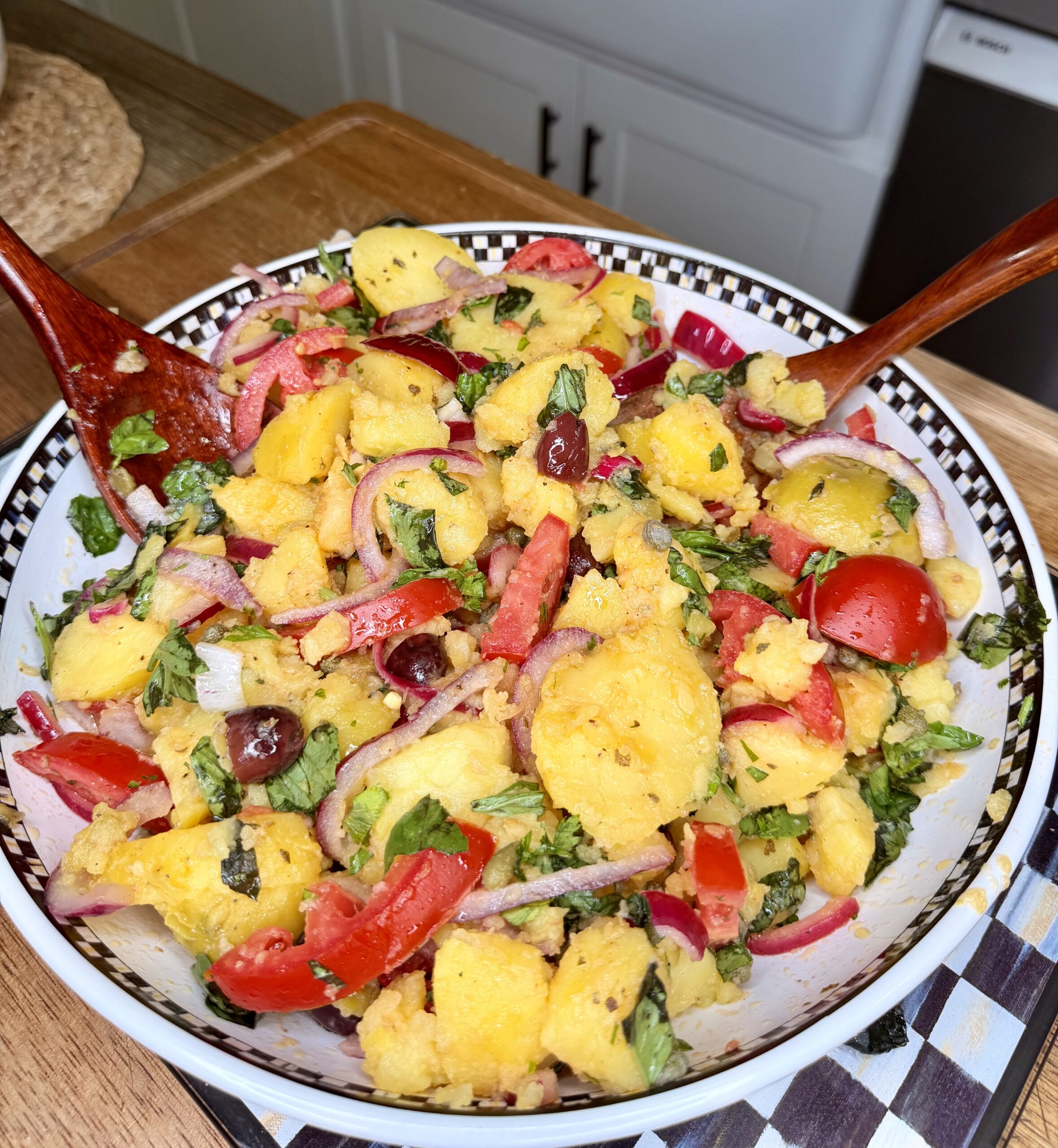 A bowl of vibrant potato salad with sliced red onions, tomatoes, olives, chopped herbs, and red peppers, mixed with a fork and spoon, set on a kitchen counter.