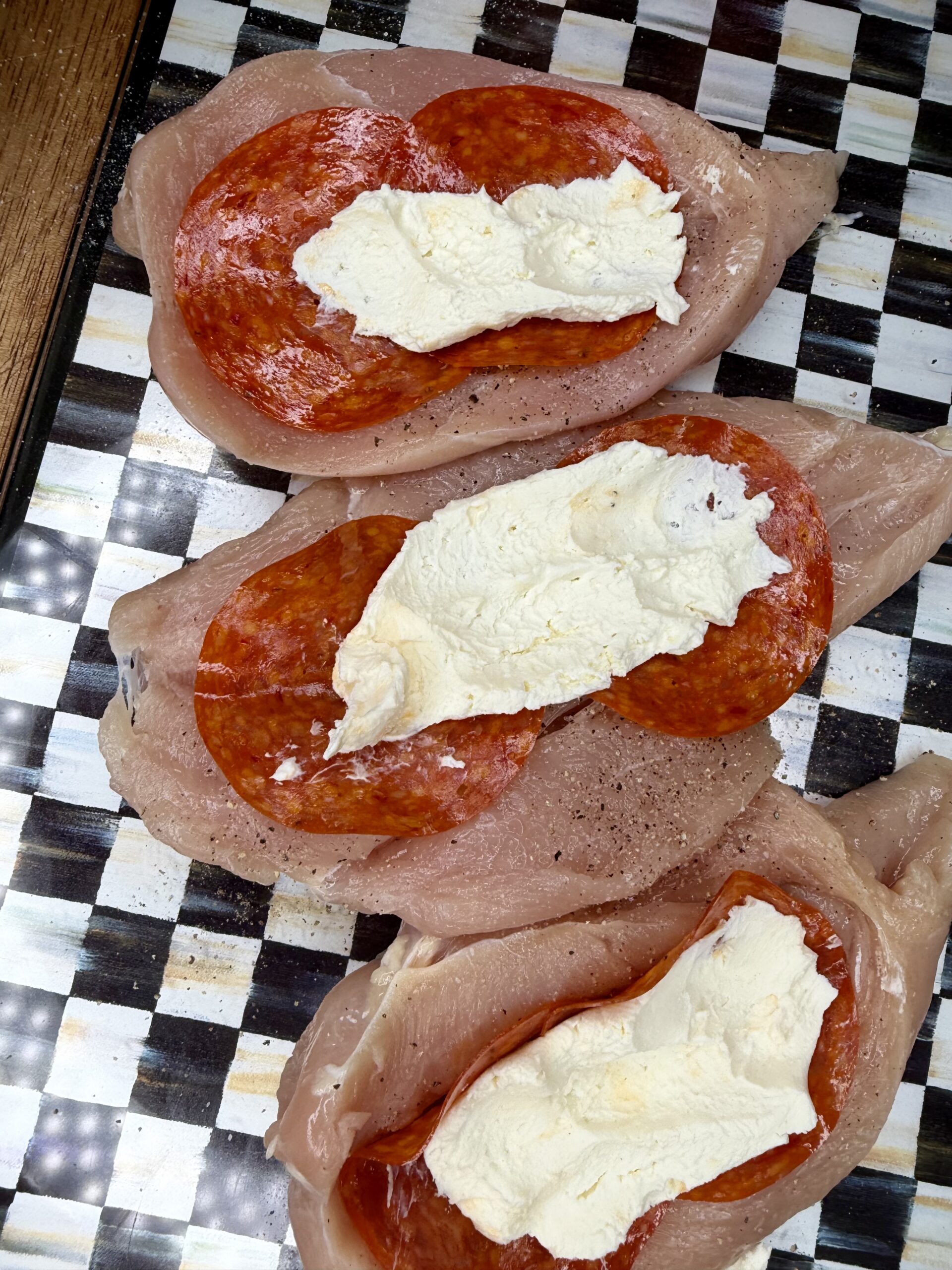 Three raw chicken breasts are topped with slices of pepperoni and a layer of creamy white cheese, arranged on a black-and-white checkered surface.