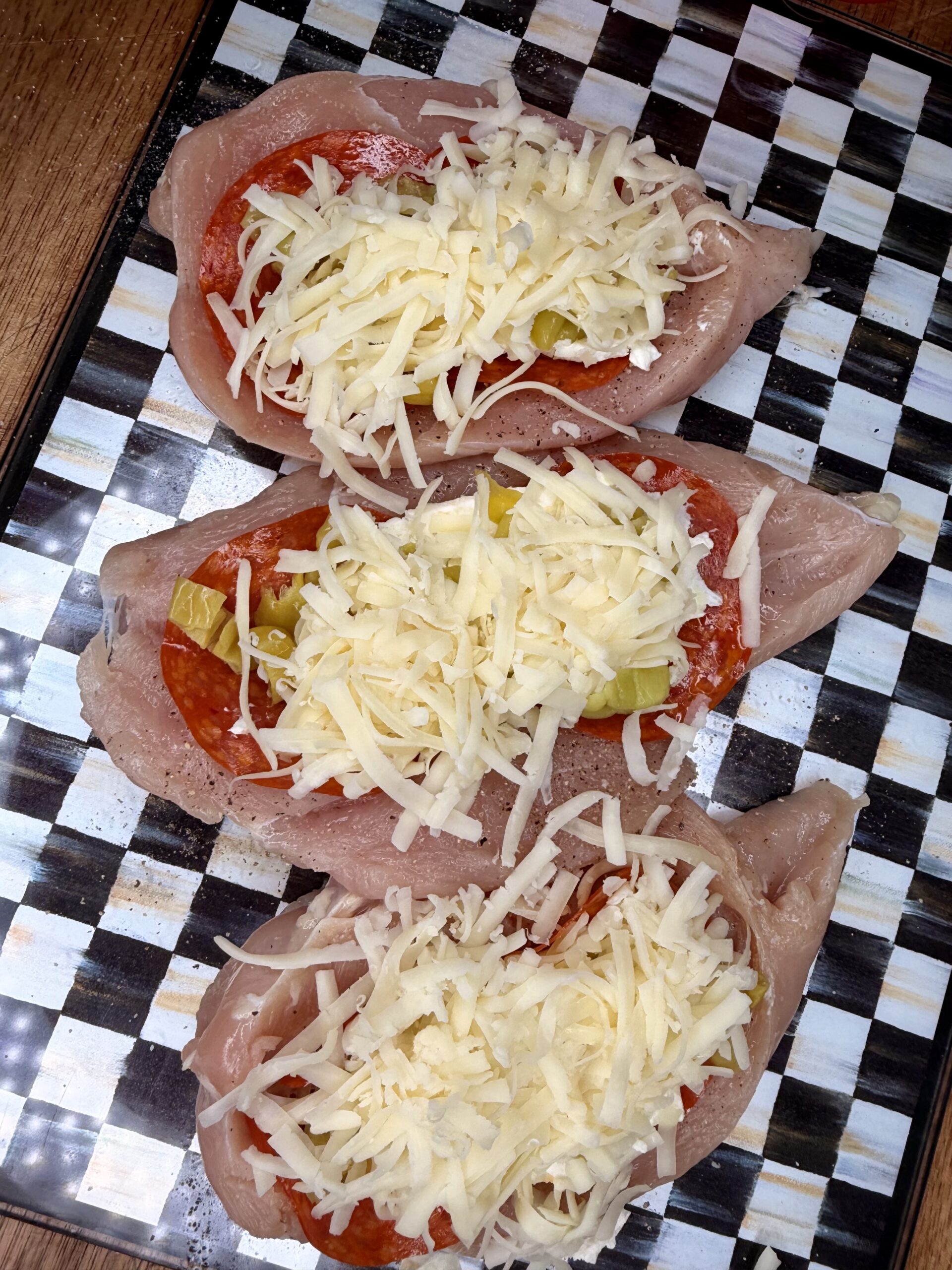 Three raw chicken breasts are sliced open and stuffed with slices of pepperoni, chopped green peppers, and shredded cheese, arranged on a black and white checkered surface.