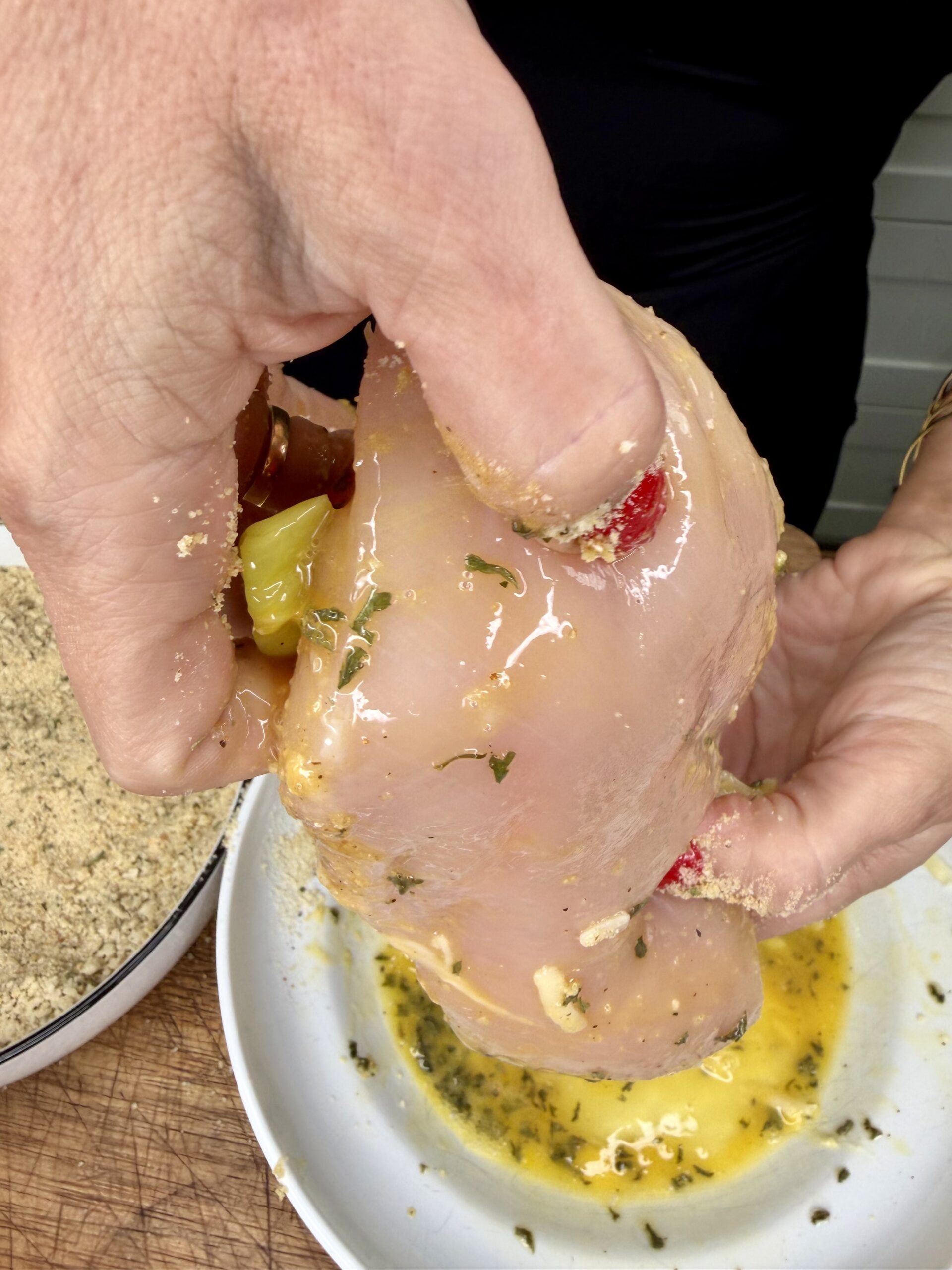A person holds a raw chicken breast coated in egg wash and herbs above a bowl, preparing it for breading; a plate of breadcrumbs is visible nearby.