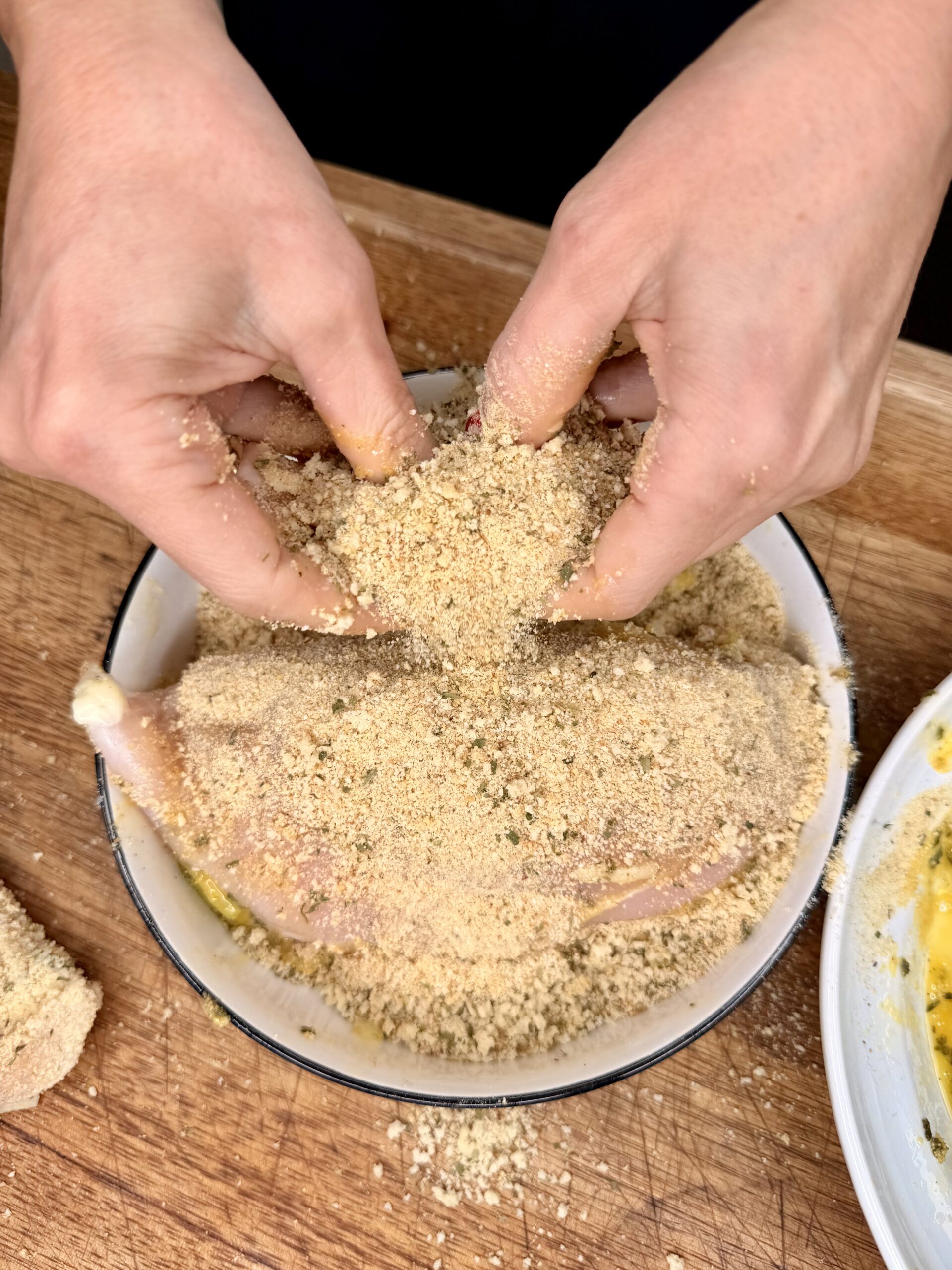 Two hands coat a raw chicken leg with seasoned breadcrumbs in a shallow bowl on a wooden surface, preparing it for cooking.