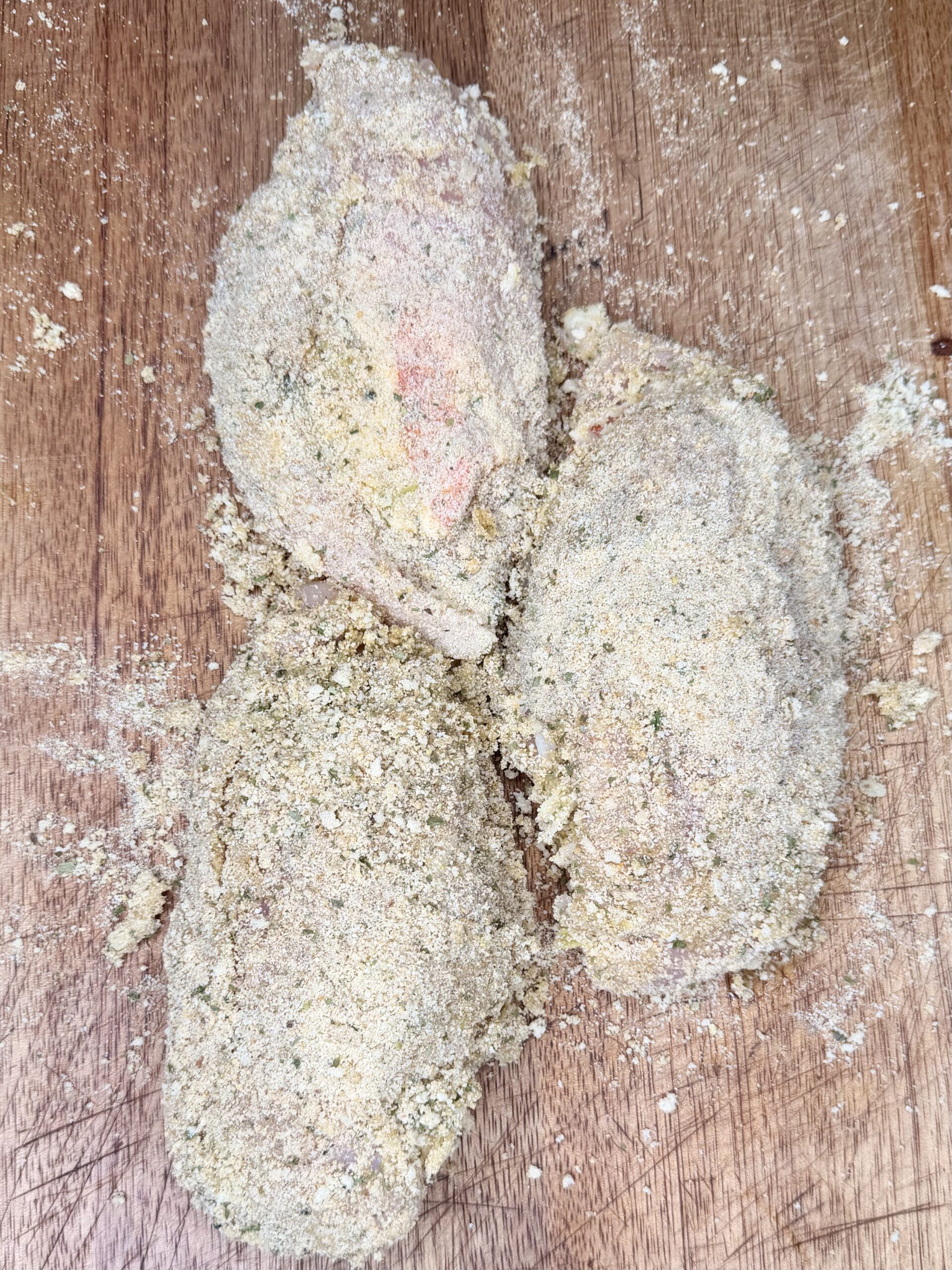 Three pieces of raw chicken coated in seasoned breadcrumbs sit on a wooden surface, ready to be cooked. Crumbs and seasoning are scattered around them.