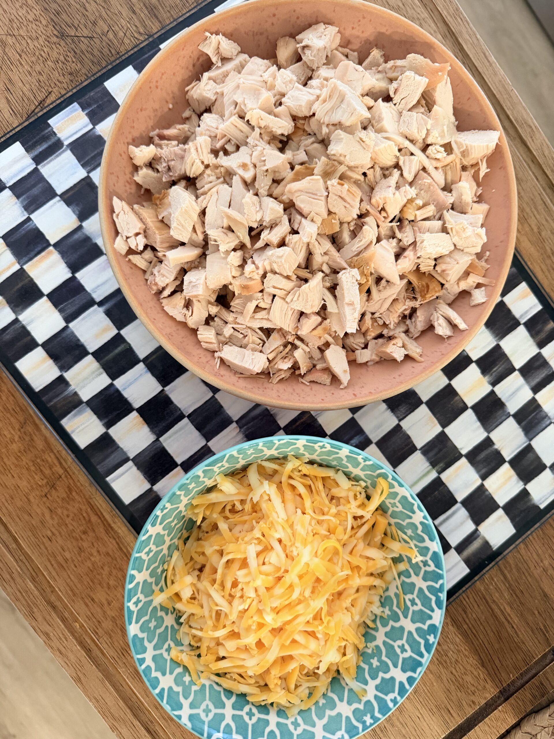 Two bowls sit on a checkered tray: one bowl contains chopped cooked chicken, and the other contains a mix of shredded yellow and white cheese. The tray rests on a wooden surface.
