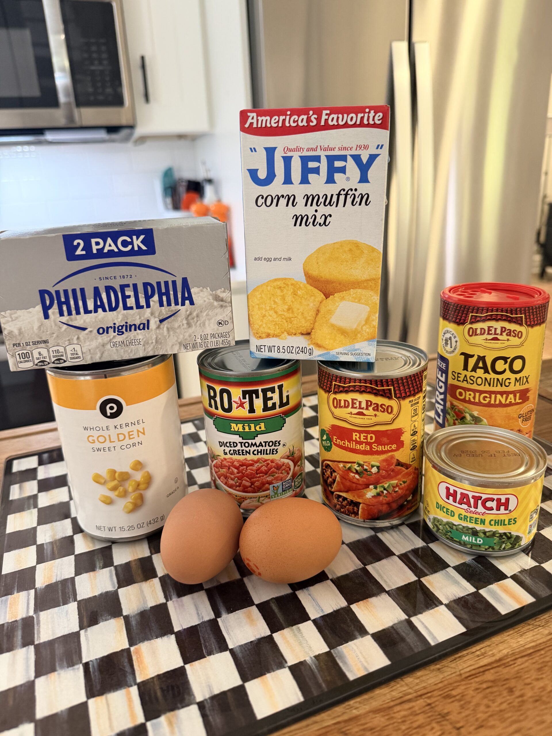 A variety of food items on a kitchen counter, including eggs, cream cheese, corn muffin mix, canned corn, diced tomatoes with green chilies, red enchilada sauce, taco seasoning, and diced green chiles.