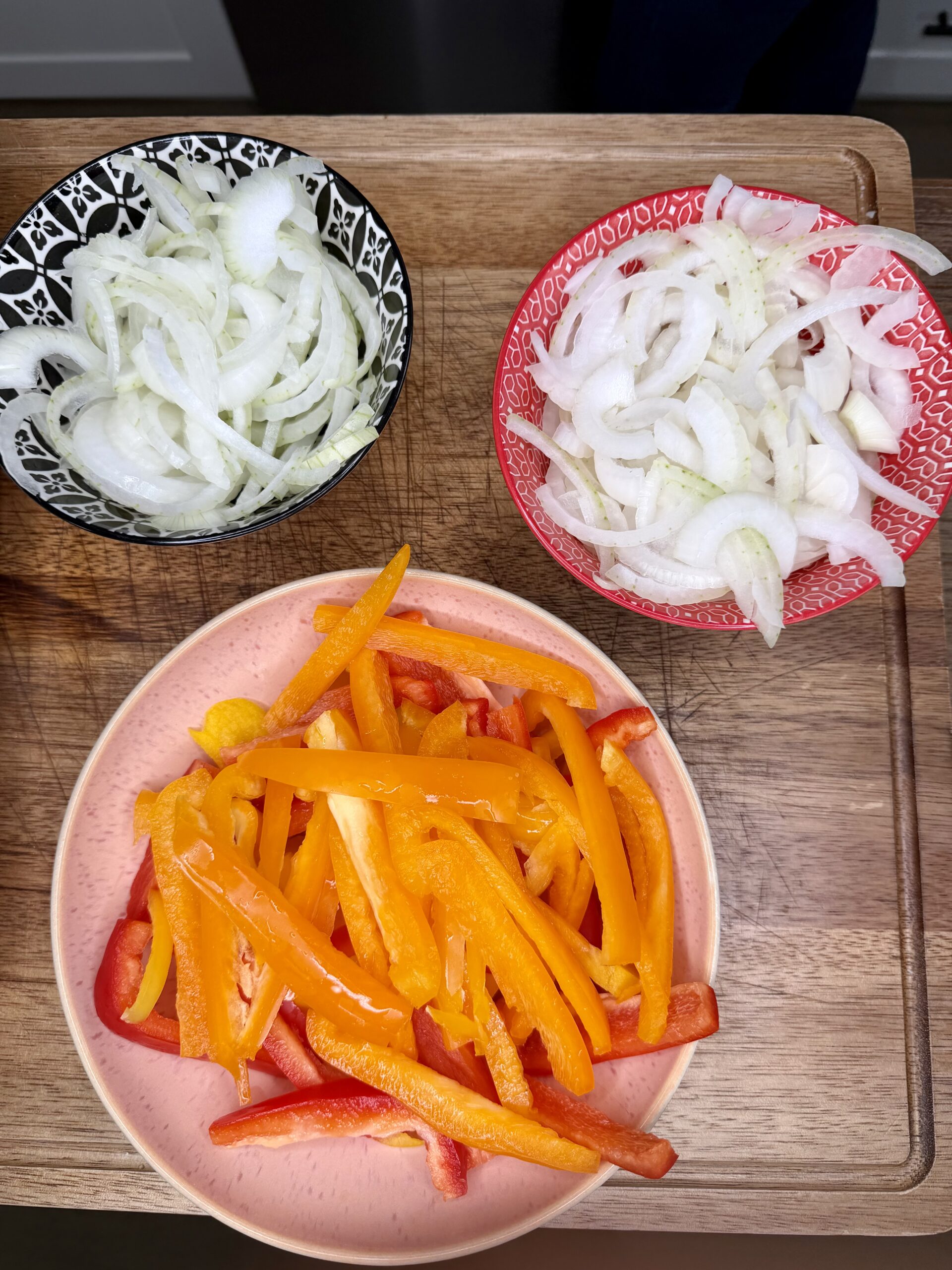 Three bowls on a wooden surface: one with sliced fennel, one with sliced onion, and one with sliced red and yellow bell peppers.